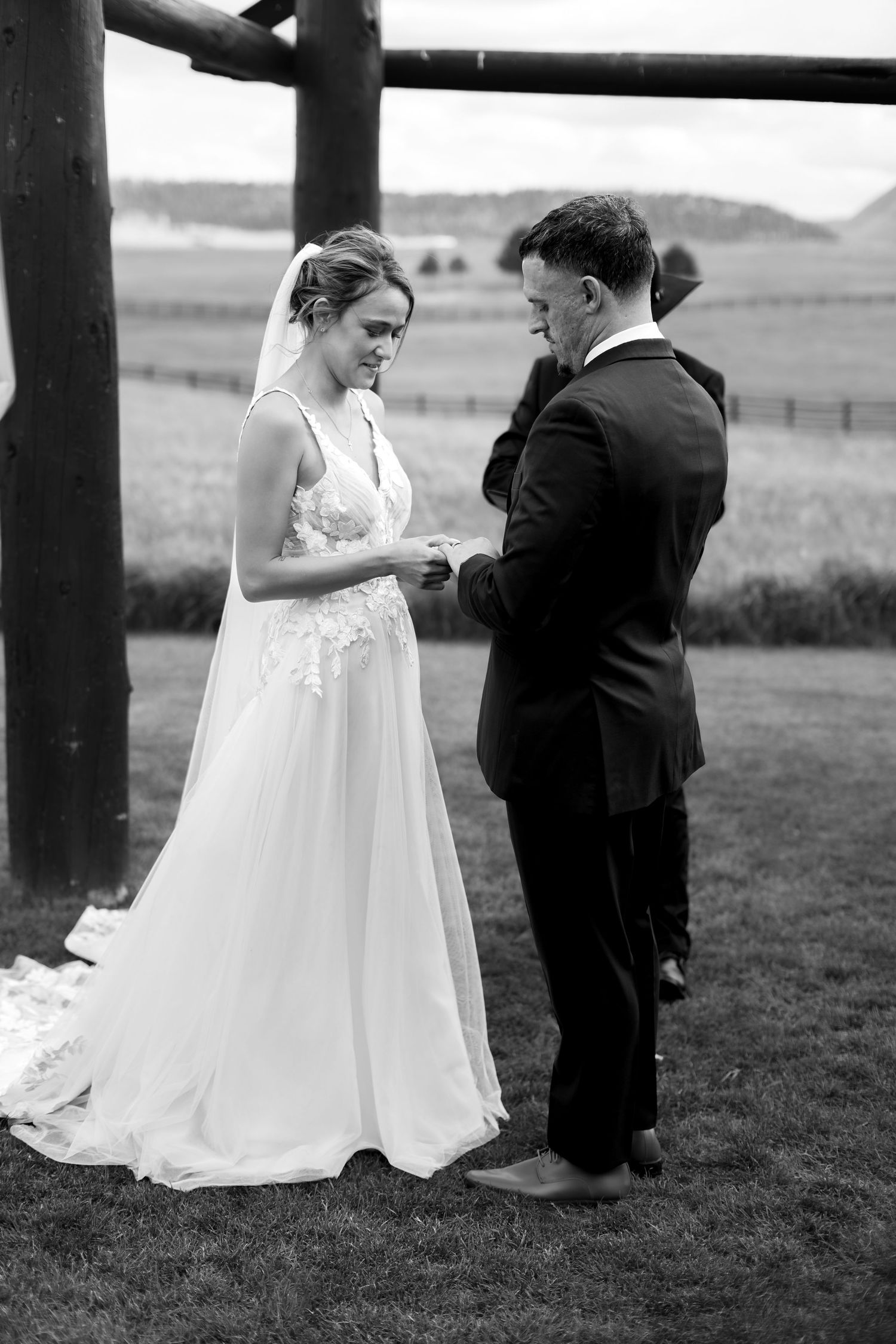 Black and white photo of bride and groom exchanging rings during an outdoor wedding ceremony on a green lawn.