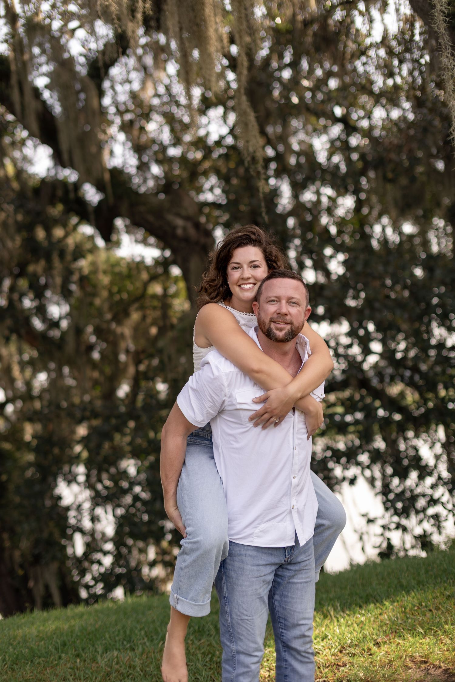 Man giving woman a piggyback ride outdoors, both smiling joyfully under a large tree.