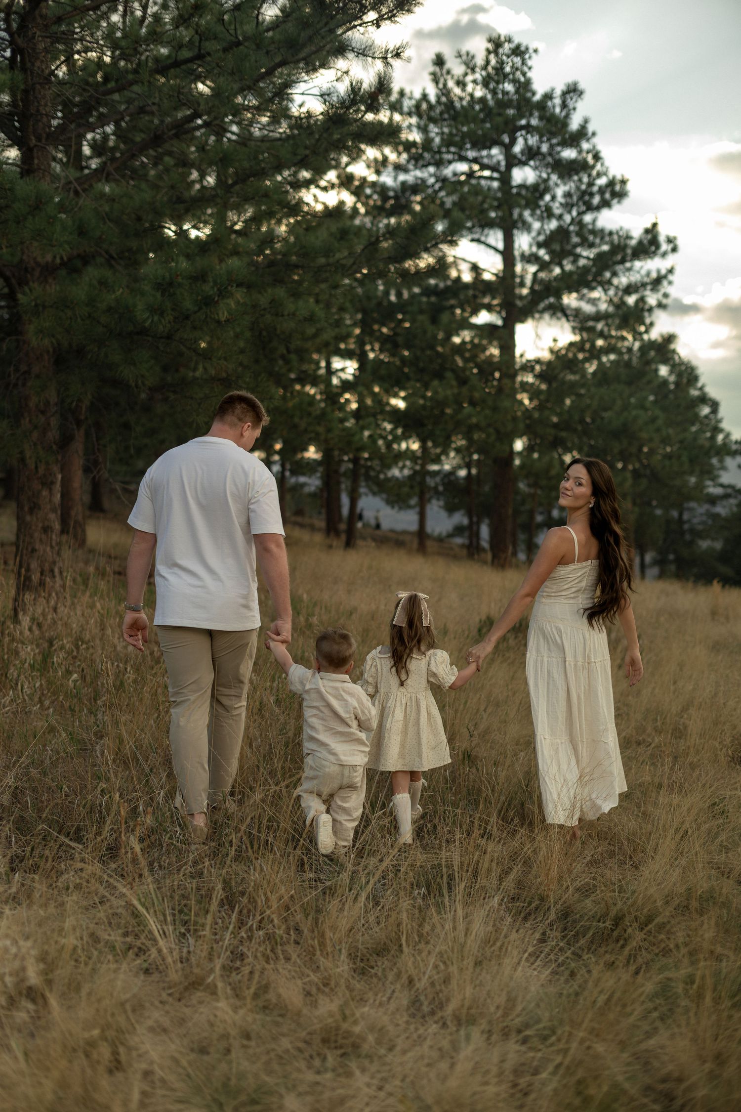 Family of four holding hands walking through golden grass field surrounded by pine trees at dusk.