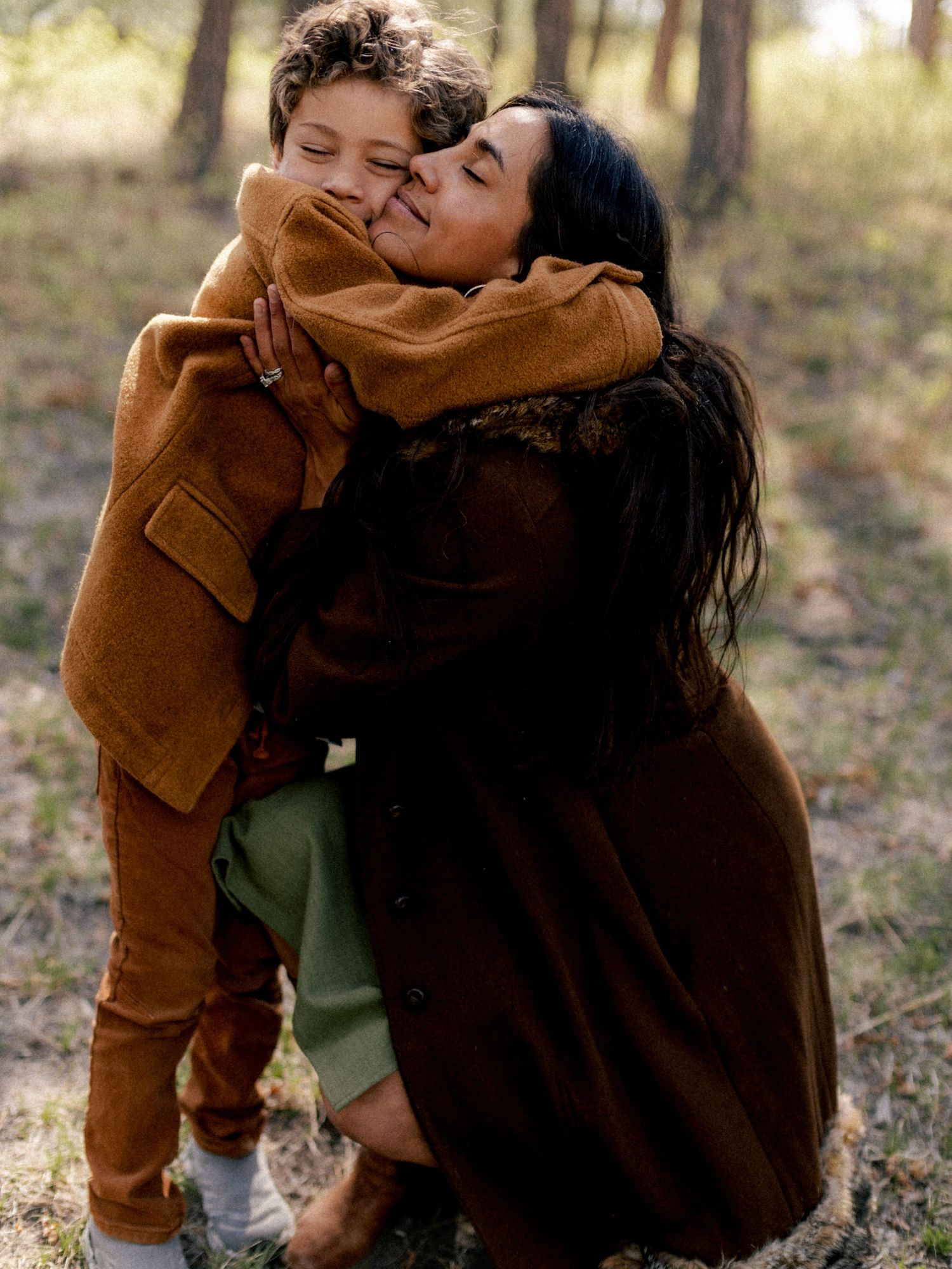 A woman in a brown coat hugs a young child in an autumn forest setting.