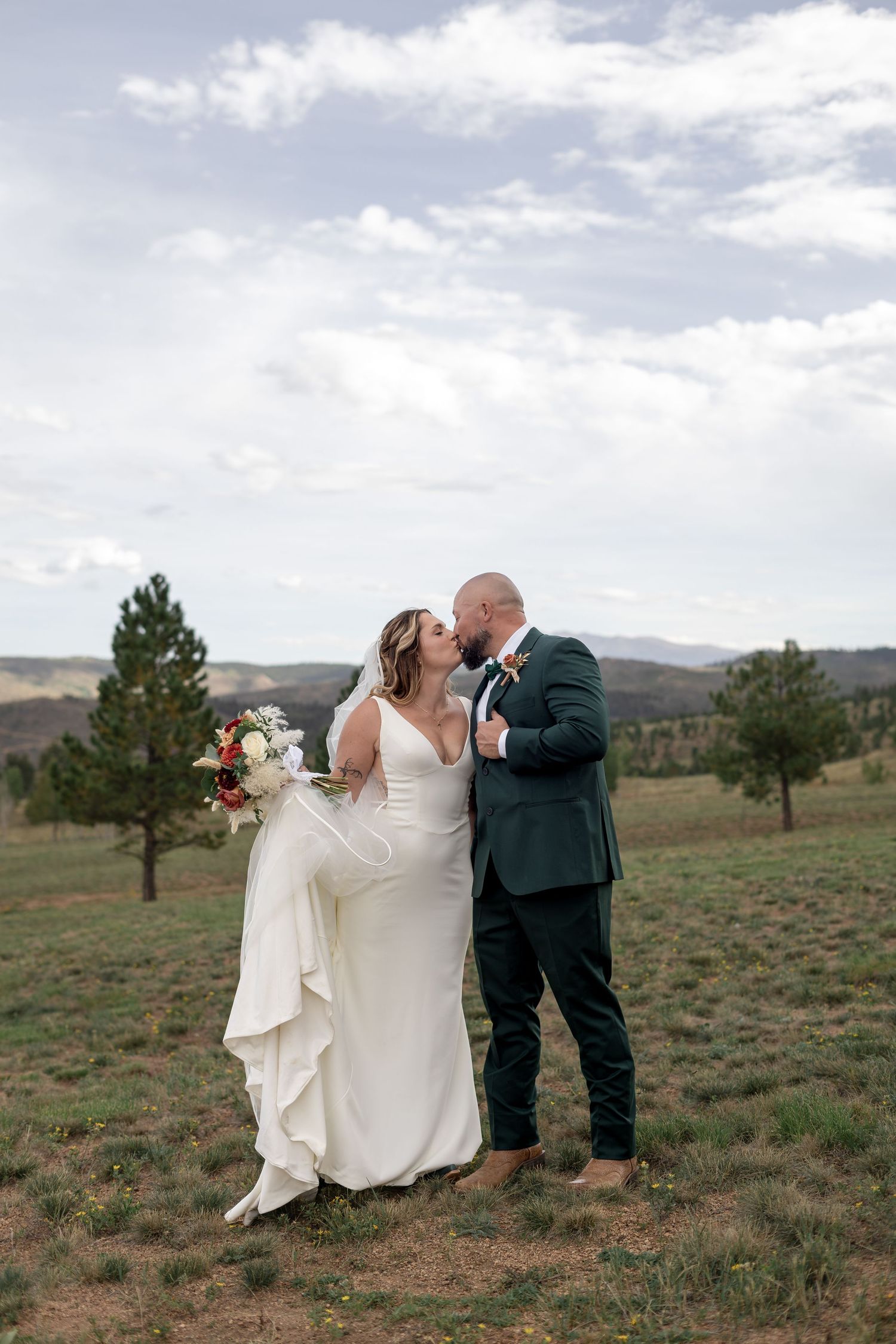 Bride and groom share a kiss outdoors in a mountain meadow, bride in white gown holding bouquet, groom in dark suit.