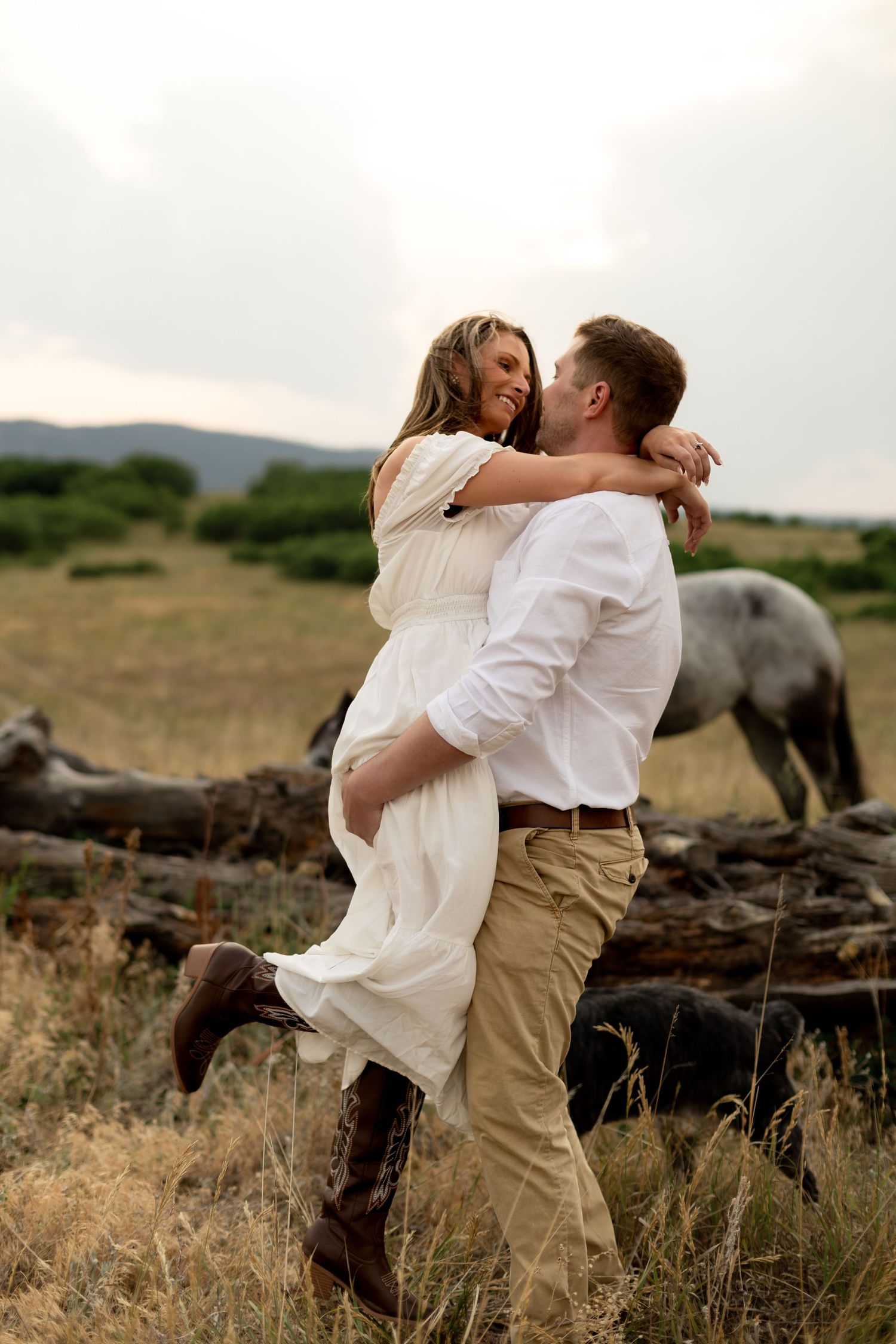 Couple embracing in a field, man lifting woman in white dress, with a horse grazing in the background.