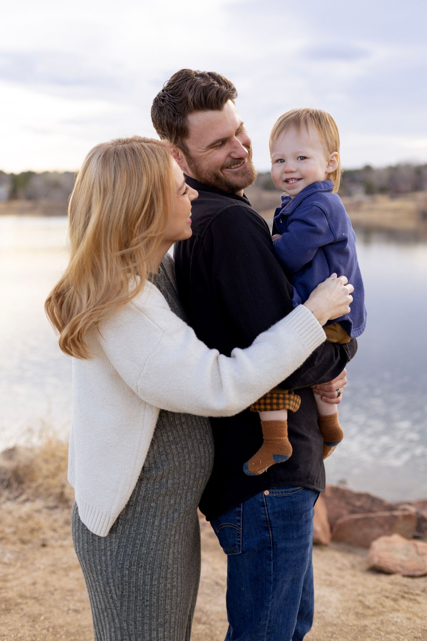 Happy family with parents holding smiling toddler boy outdoors near a lake on a cloudy day.