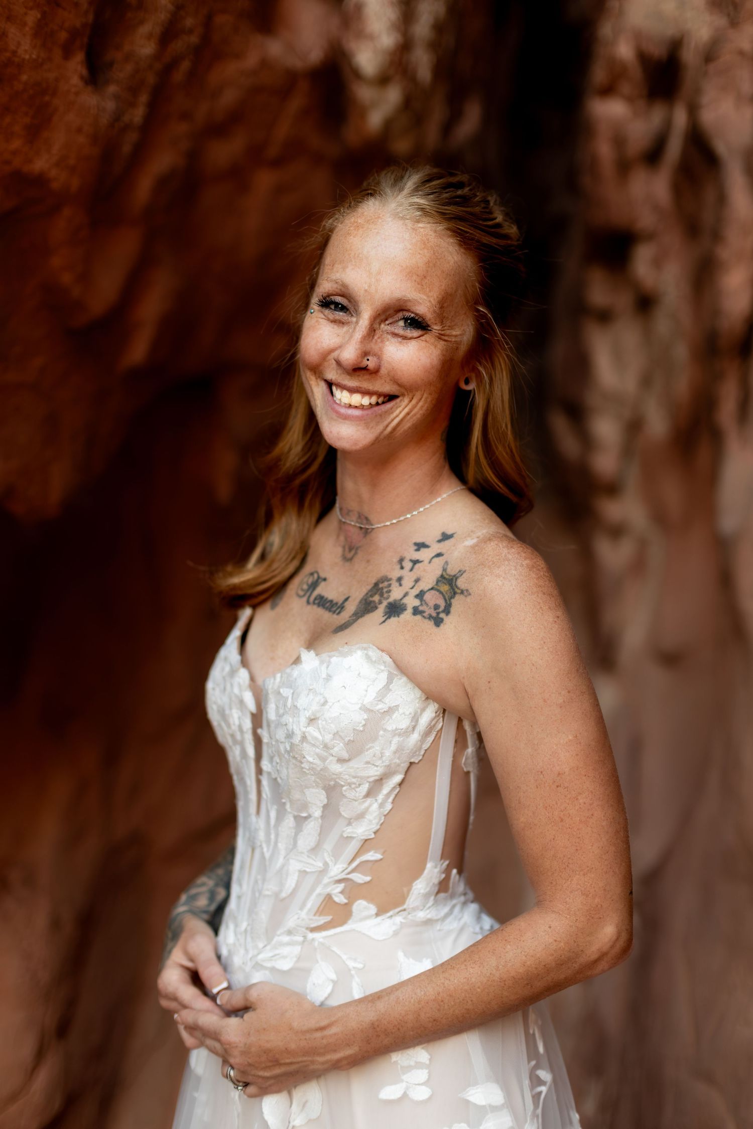 Smiling bride in floral lace wedding gown with shoulder tattoo poses in red rock canyon setting.