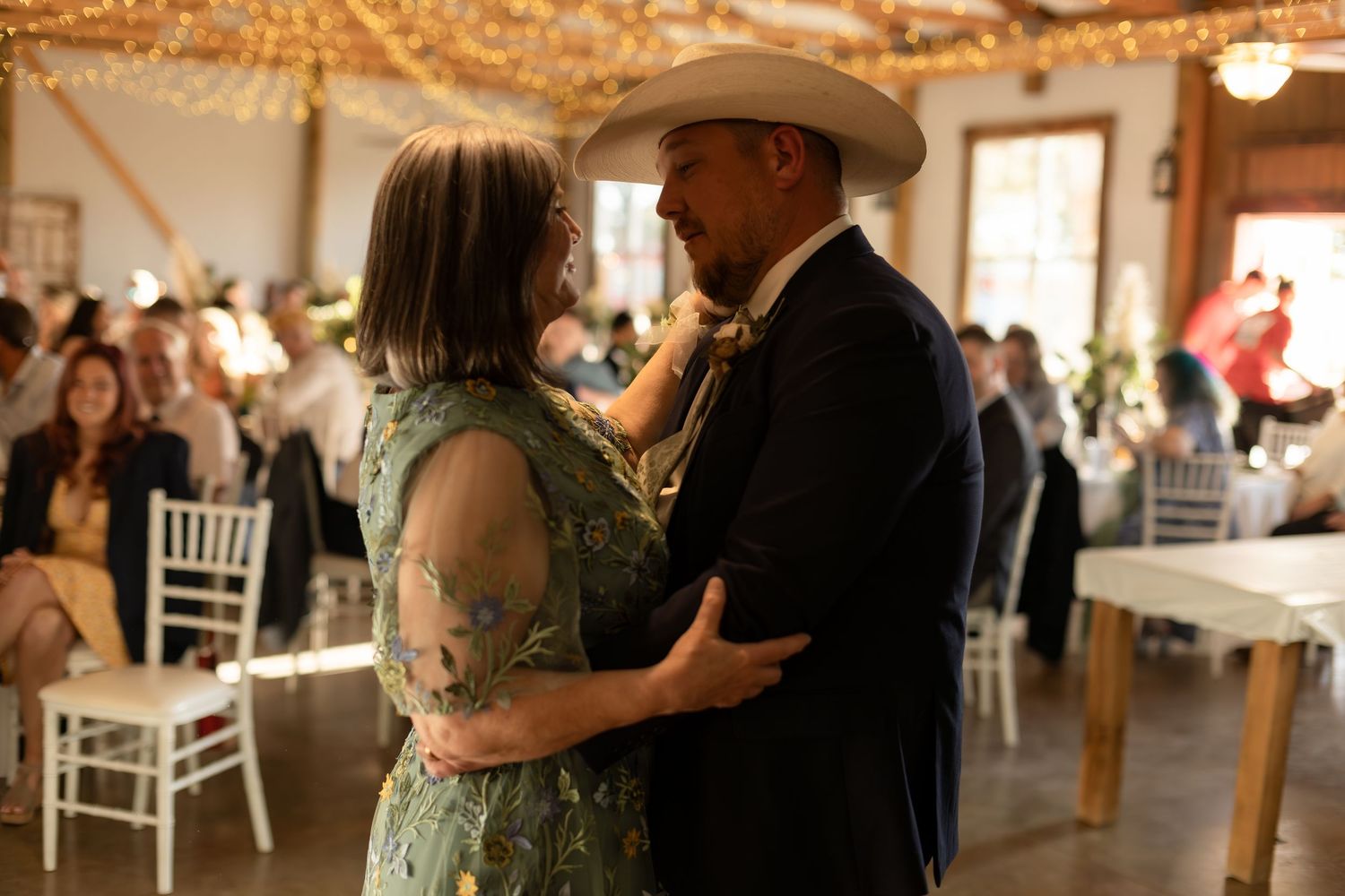 Groom in cowboy hat and navy suit shares mother-son dance in warmly lit rustic barn reception hall.