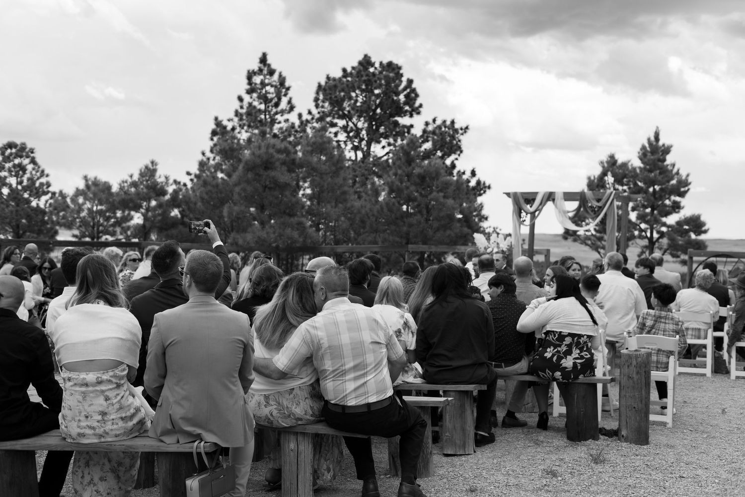 Black and white photo of wedding guests seated on wooden benches at an outdoor ceremony with trees in background.