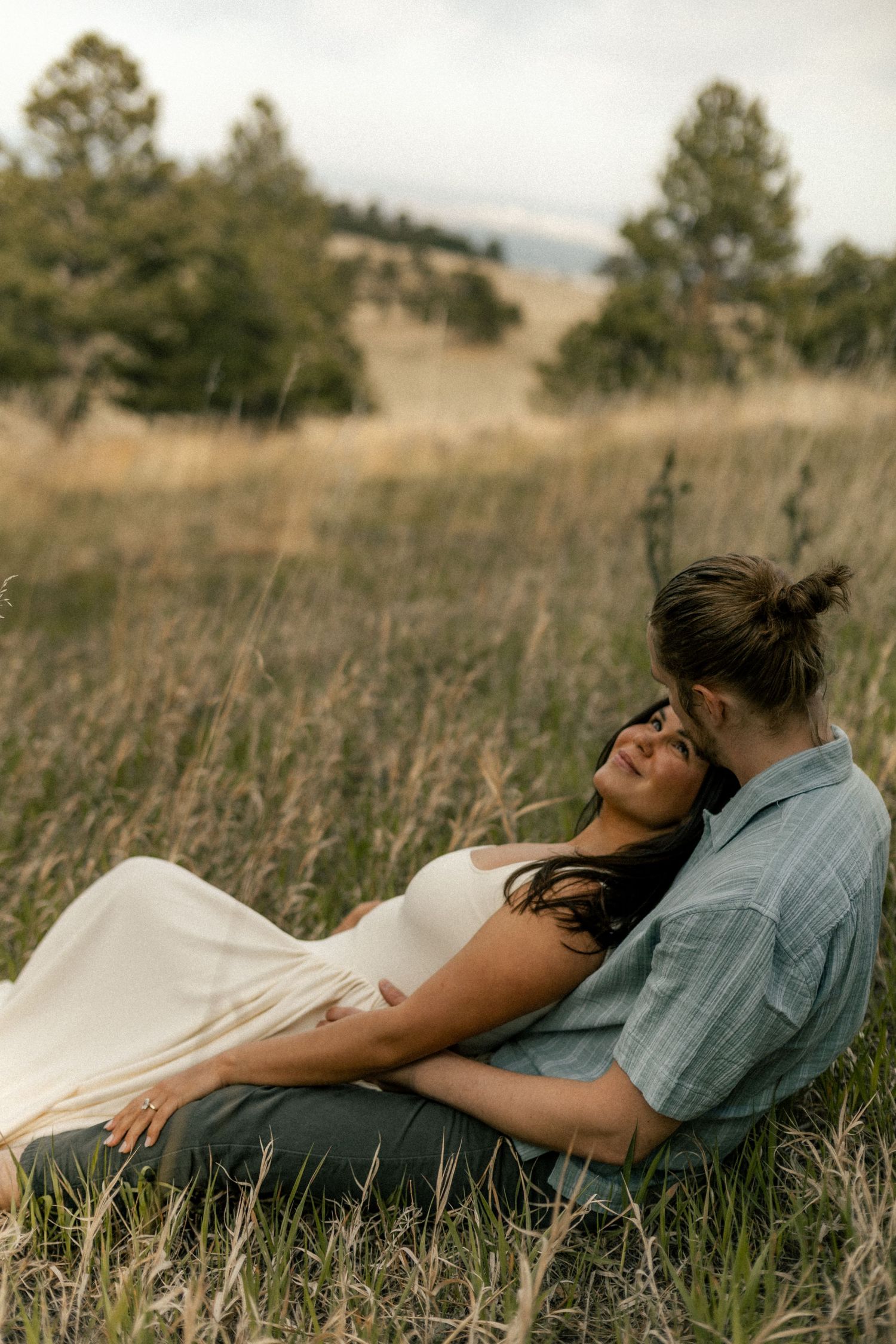 Couple relaxing in tall grass field, woman leaning back on man in blue shirt, serene outdoor setting.
