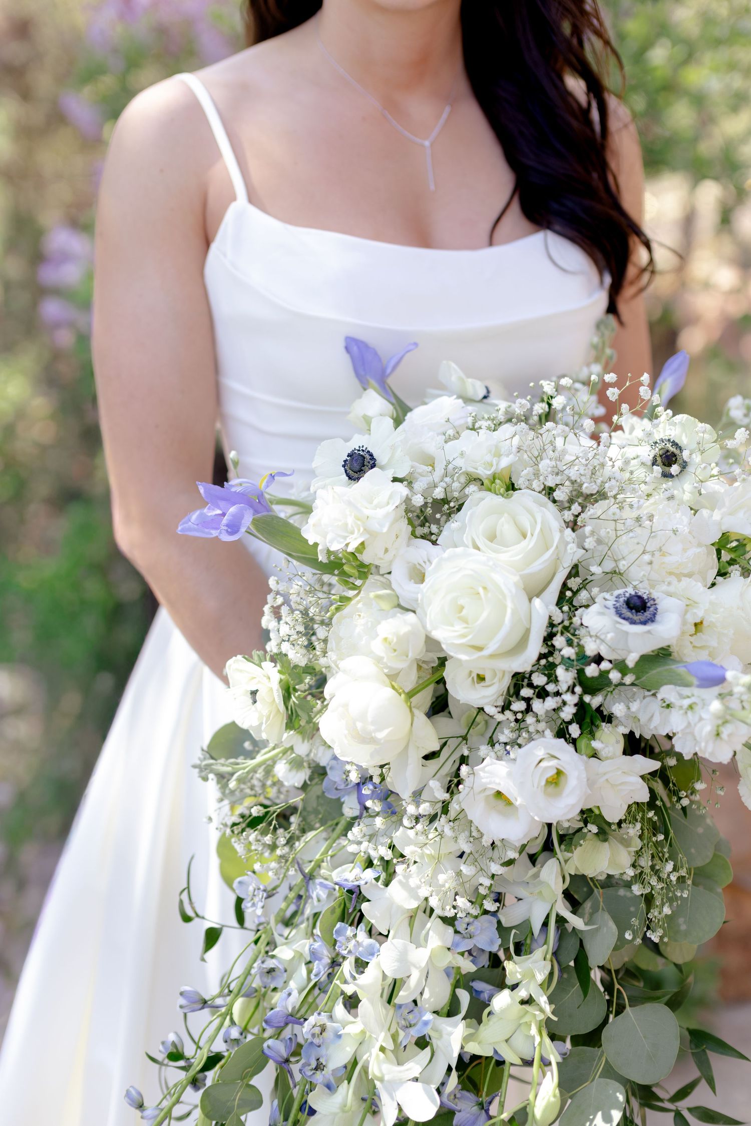 Bride in white spaghetti strap gown holding lush cascading bouquet of white roses, anemones, and blue flowers.