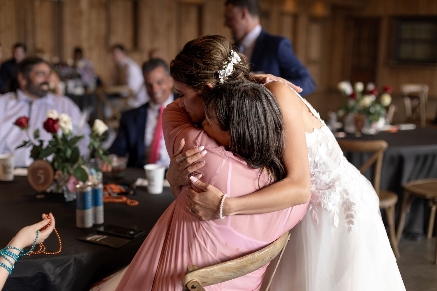 Bride in white lace gown embraces guest in pink dress during wedding reception in rustic barn.
