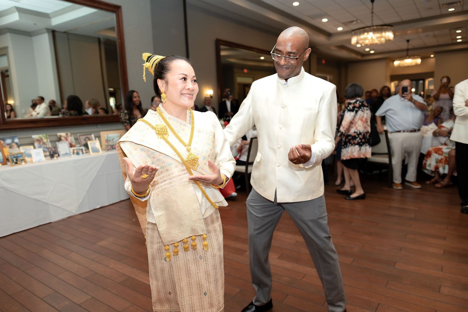 Couple dancing at elegant reception, woman in traditional gold Lao dress and man in white shirt, ballroom setting.