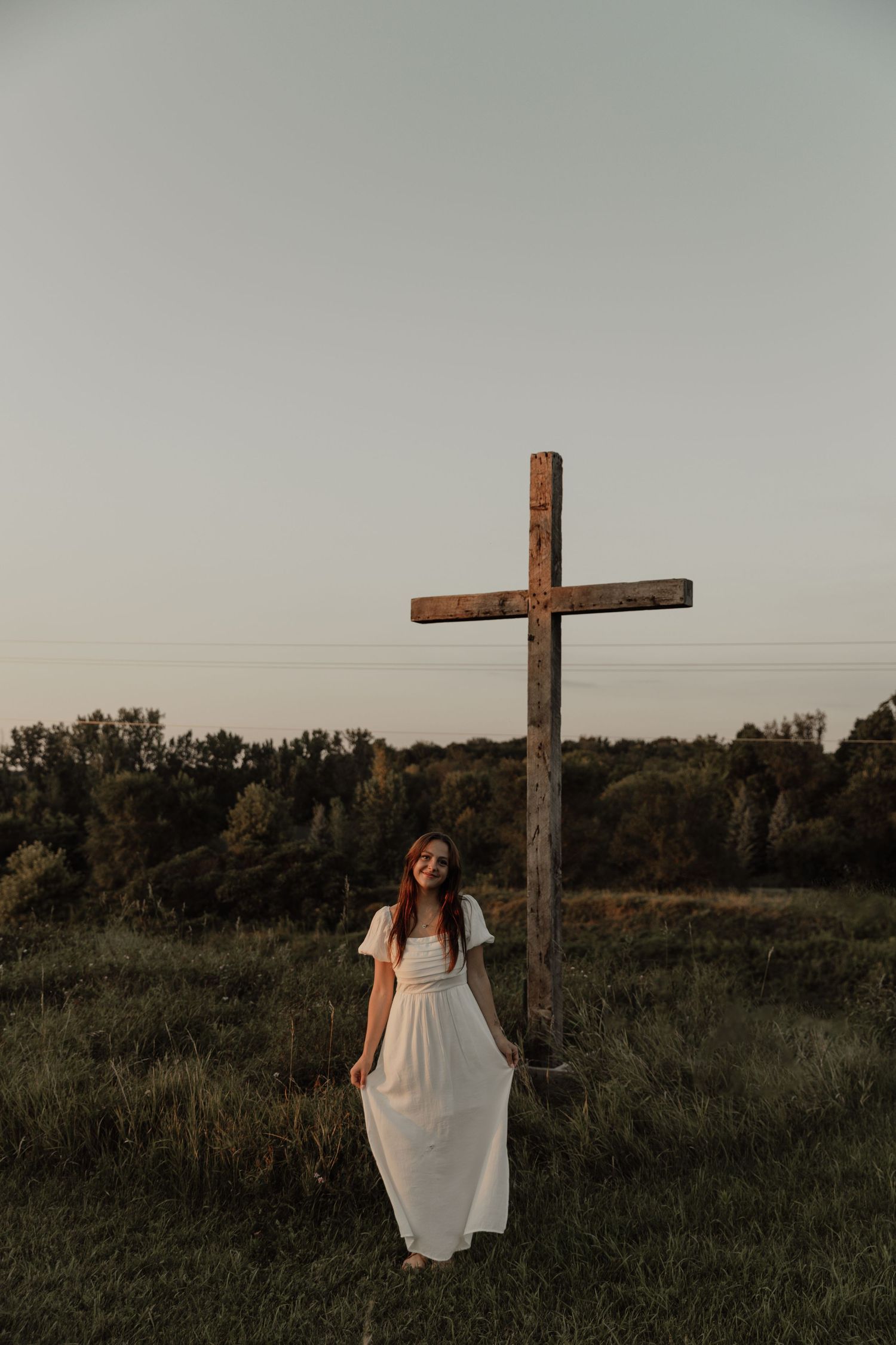 White dress portrait beside wooden cross in field during golden hour.
