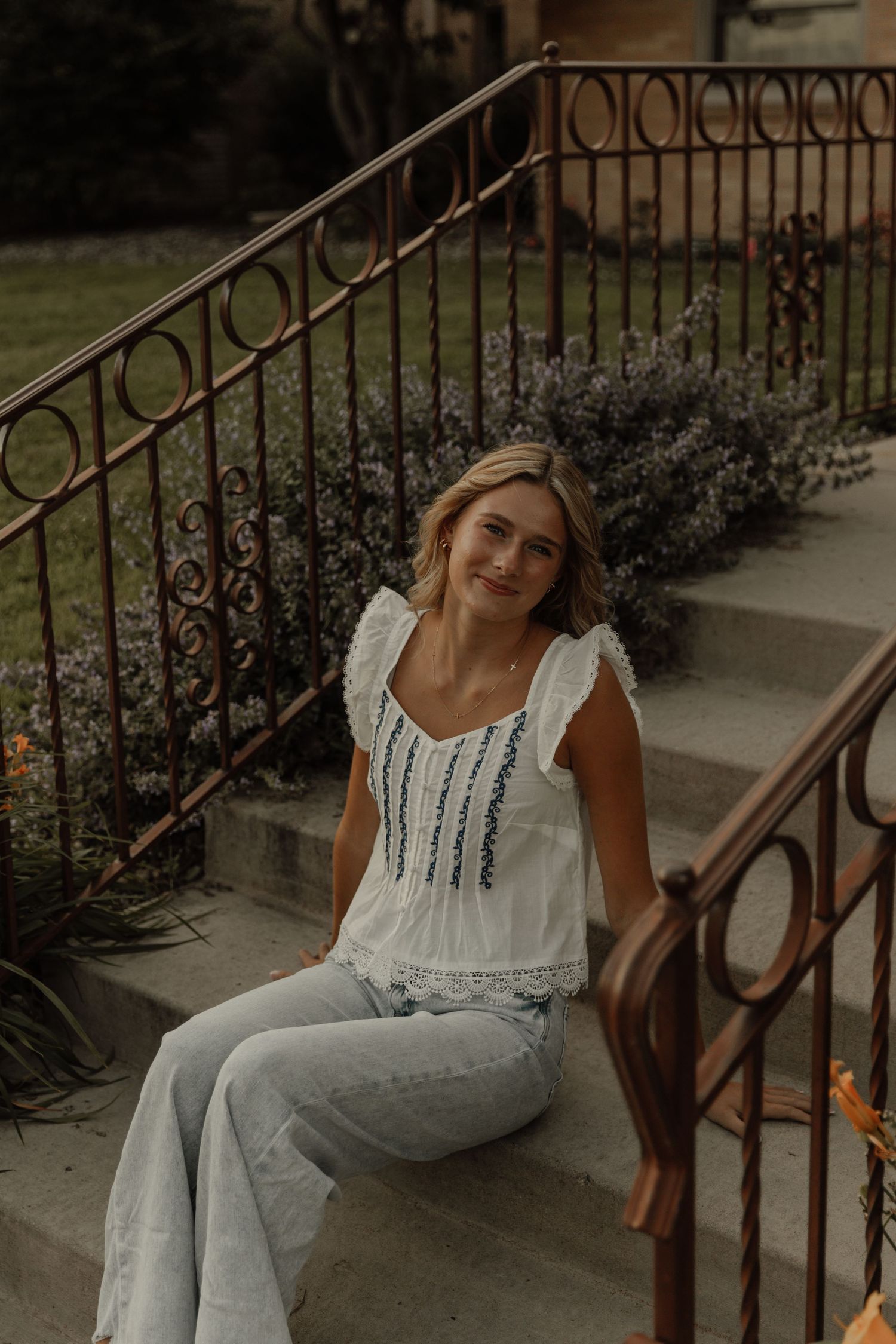 A person in a white eyelet dress poses casually on rustic outdoor steps with ornate metal railings.
