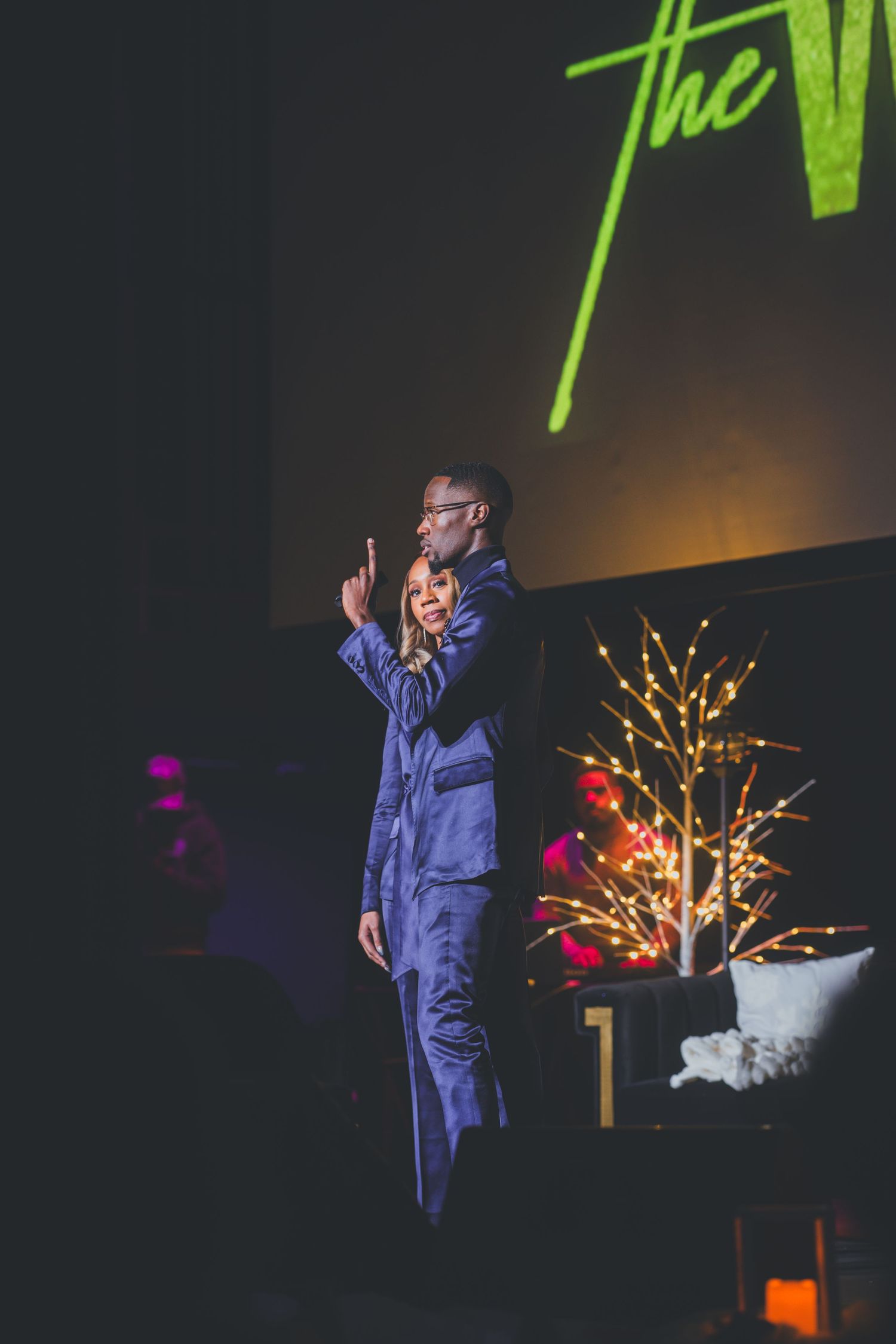 Performer in blue attire gestures dramatically on a dimly lit stage with green accents.