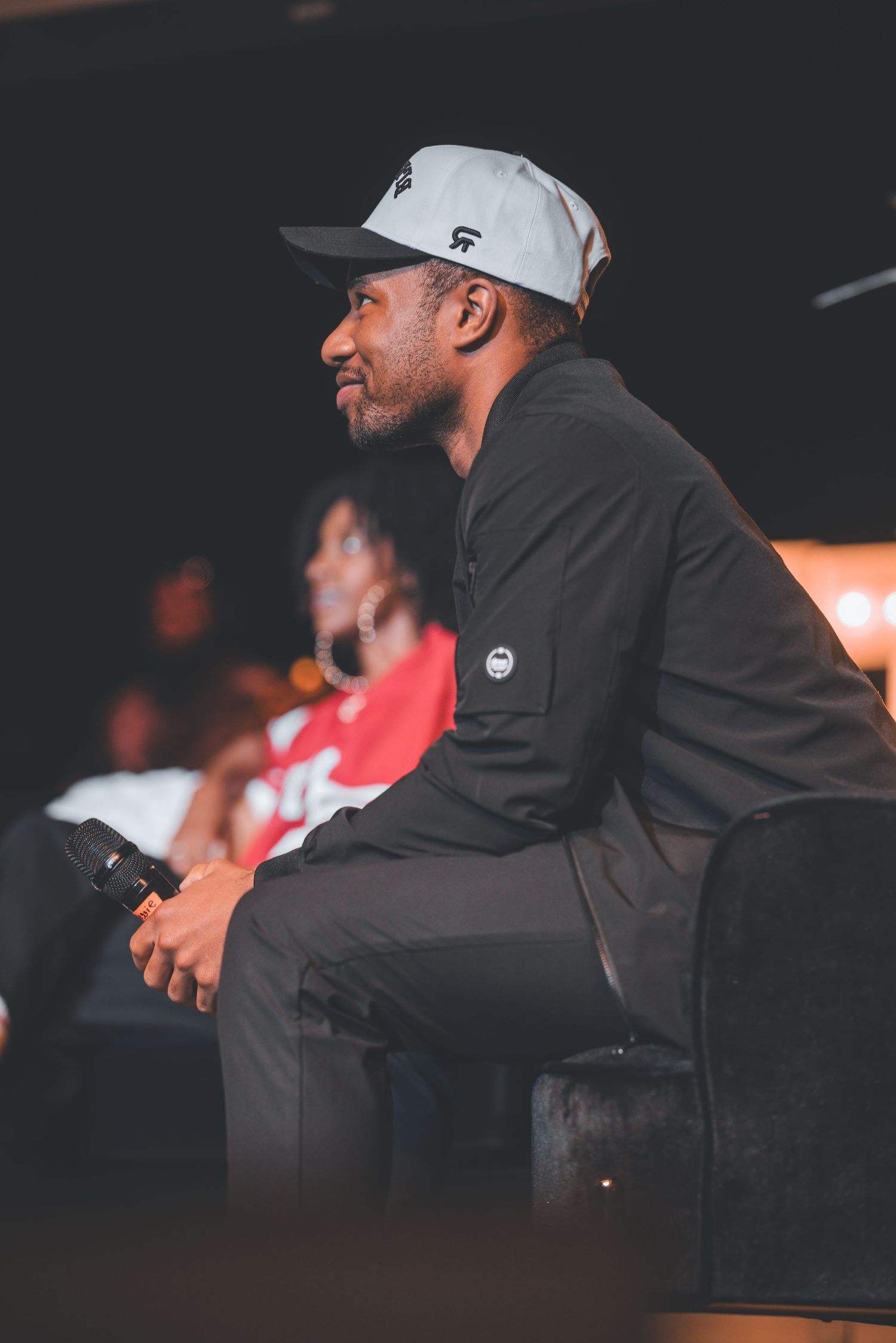 Person in a gray cap and black jacket sits on stage during an event.