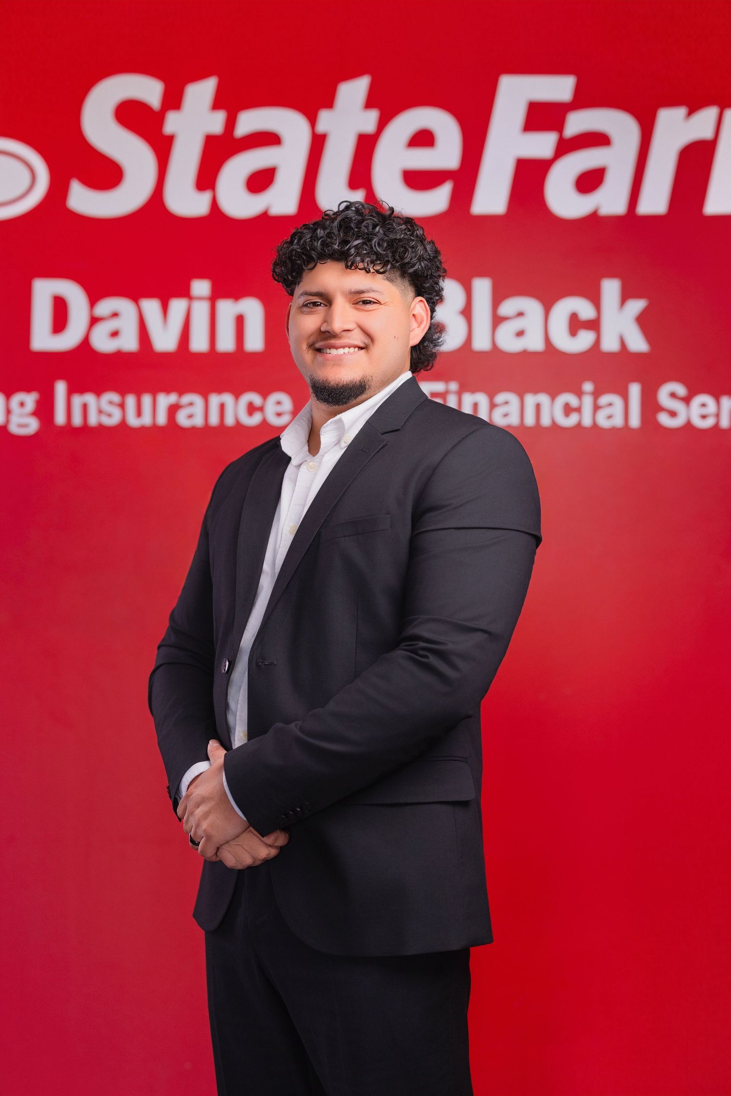 Professional portrait against a red State Farm backdrop showing person in black suit smiling.