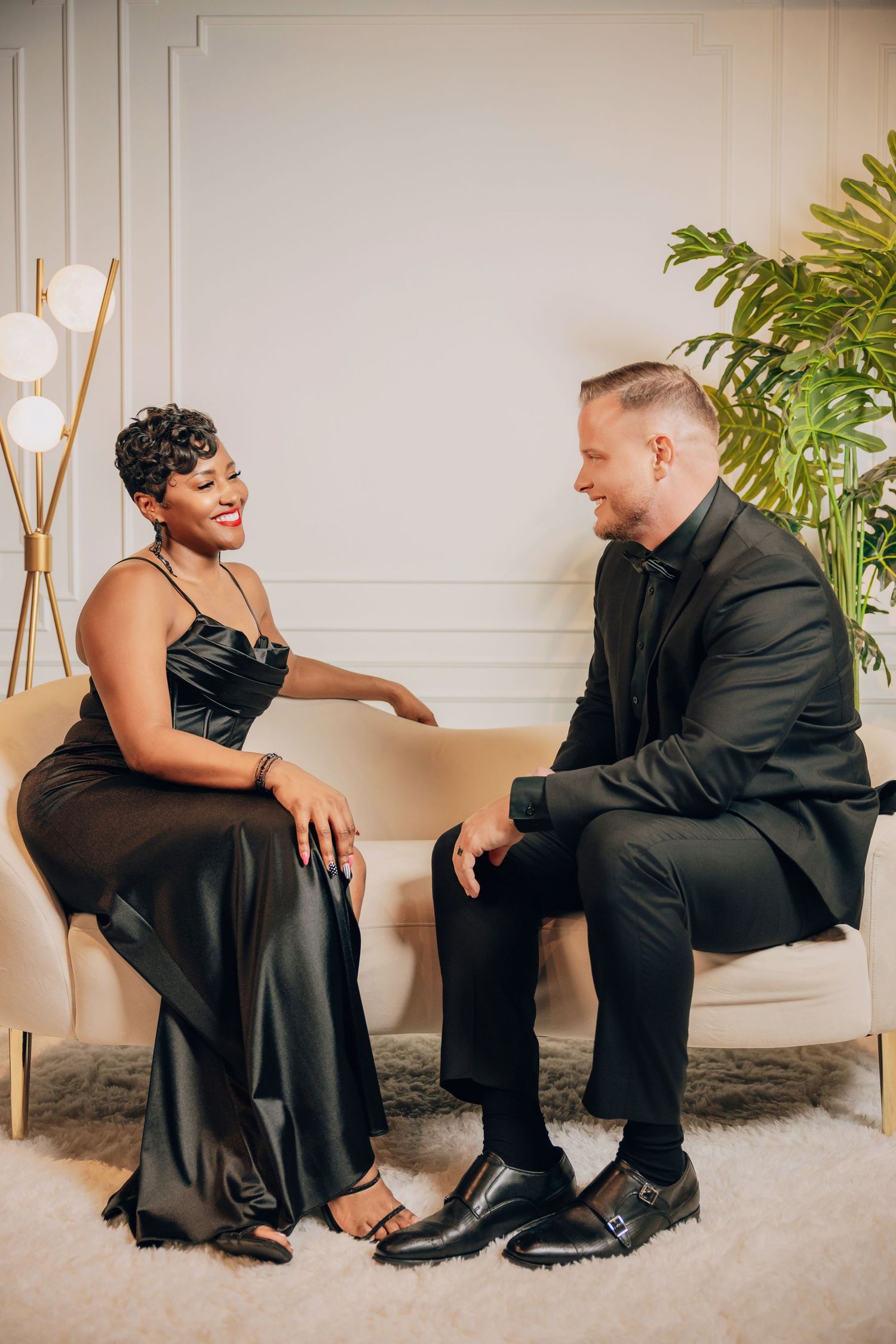 A couple wearing black formal attire shares intimate conversations while sitting on a white couch in a studio setting.