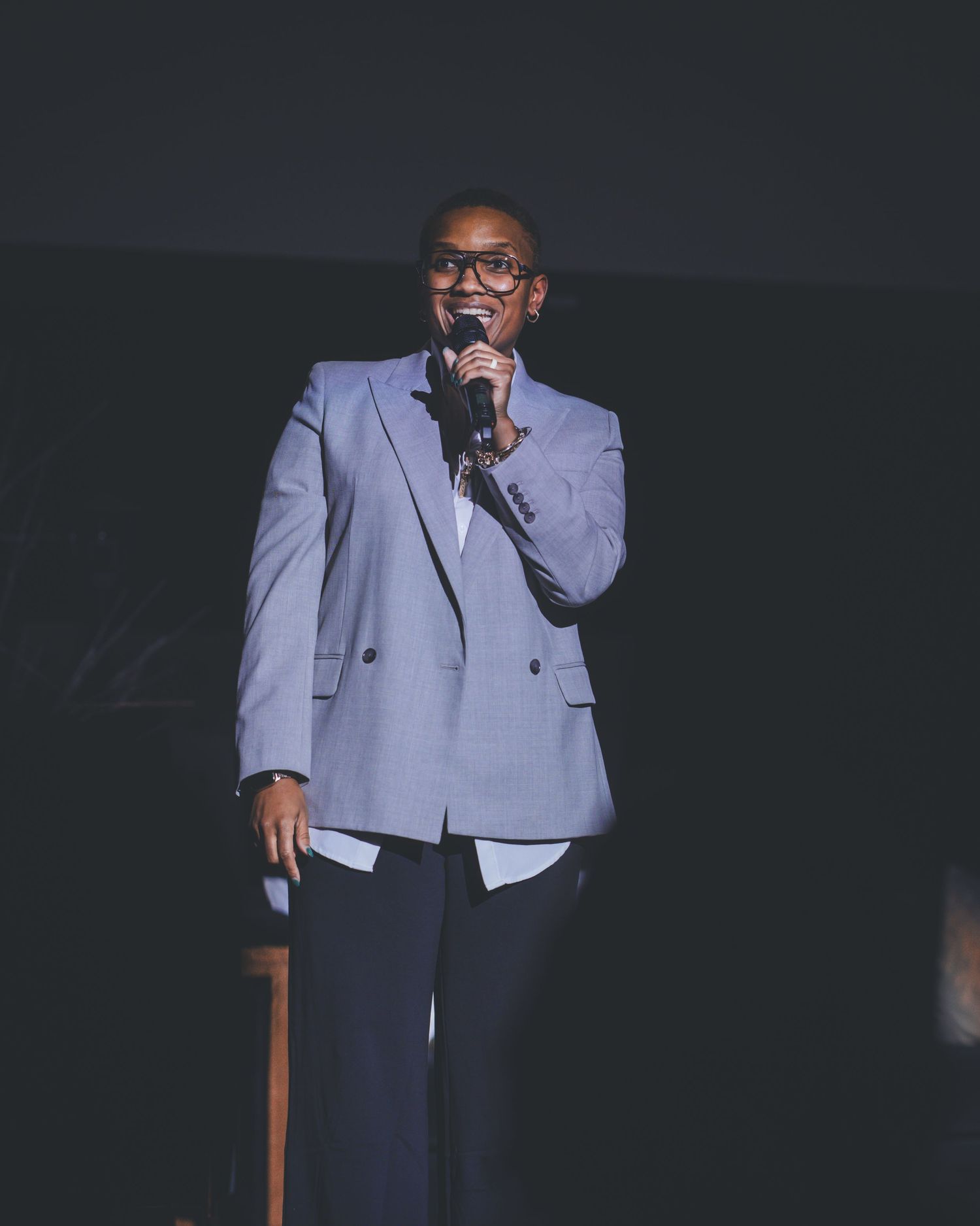 Performer in light gray suit jacket speaks into microphone on dark stage.