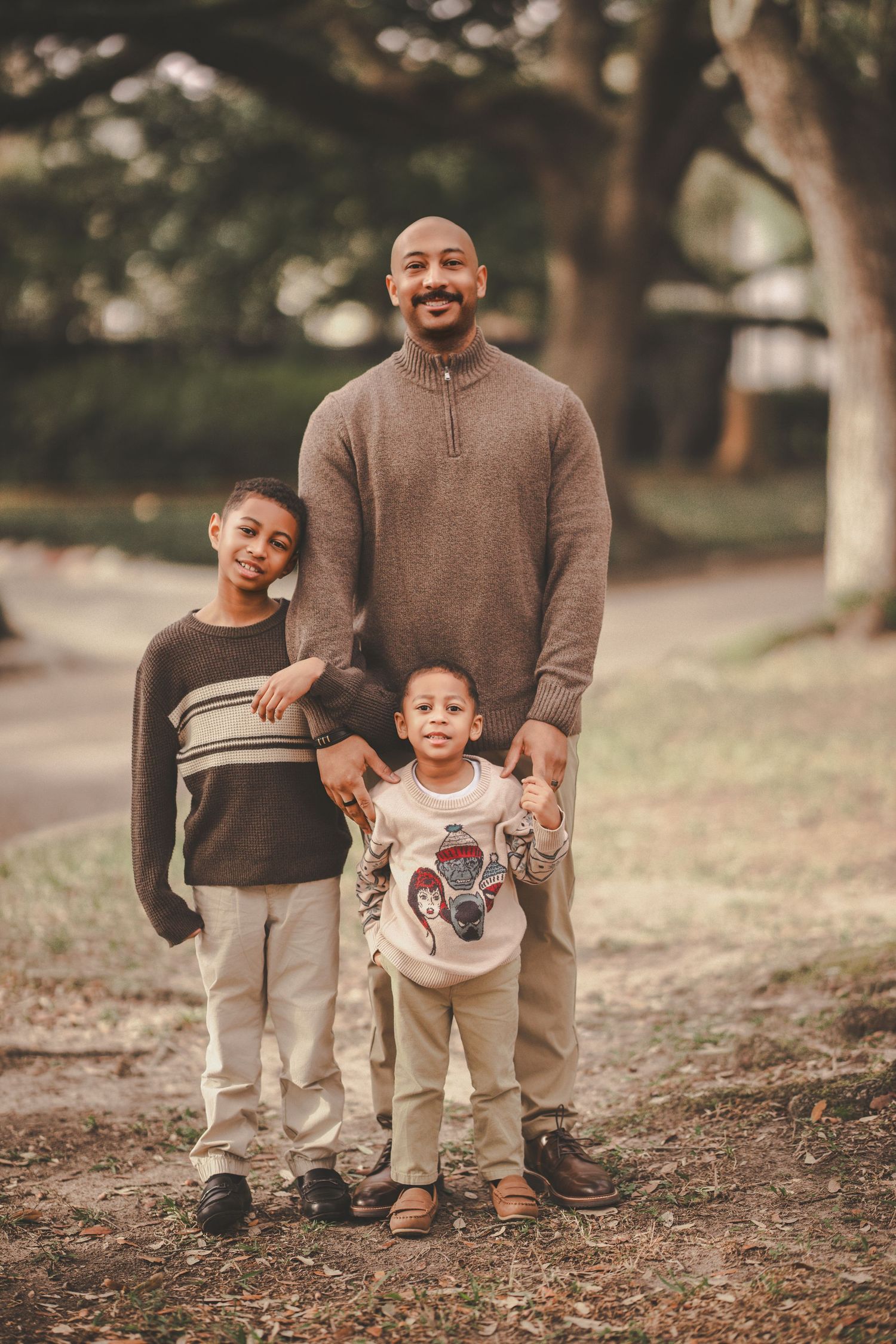 A parent and child wearing matching brown outfits pose playfully outdoors on a wooded path during autumn.
