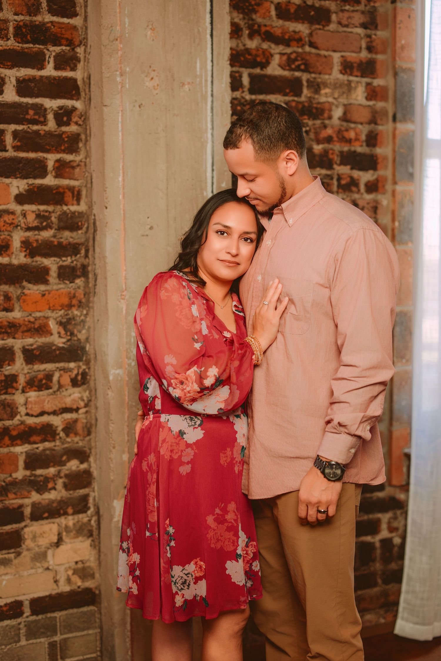 A couple in romantic poses against a brick wall, with her wearing a red floral dress and him in a beige shirt.