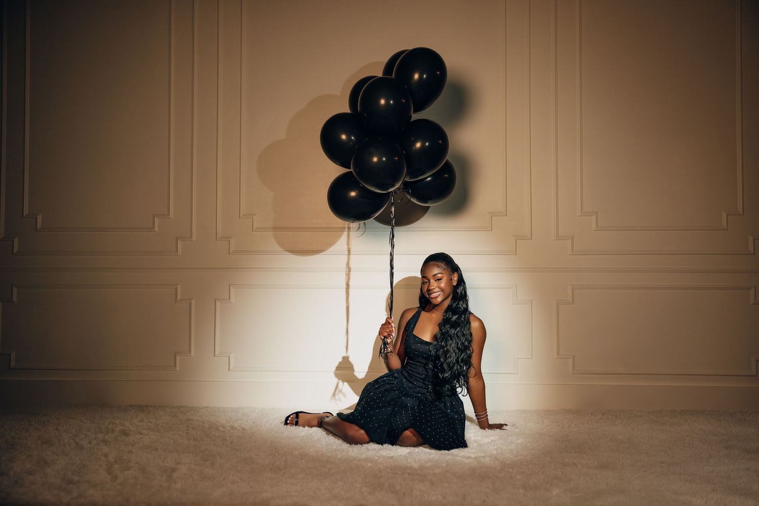 A person in an elegant black dress sits on the floor holding a bundle of black balloons against a light colored wall.