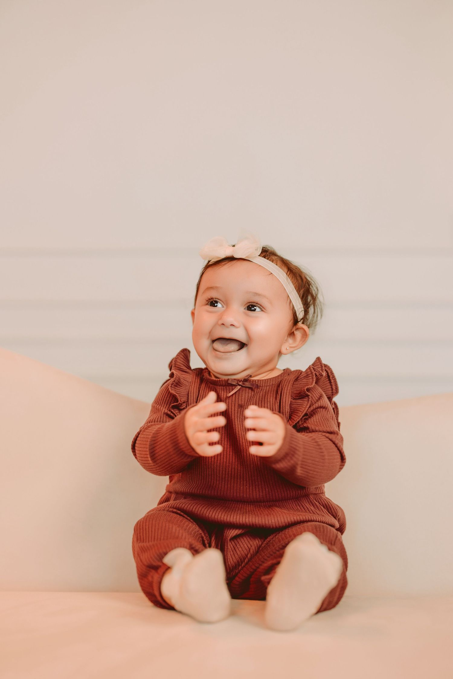 Baby wearing brown knit sweater sits and smiles on light beige background in studio setting.