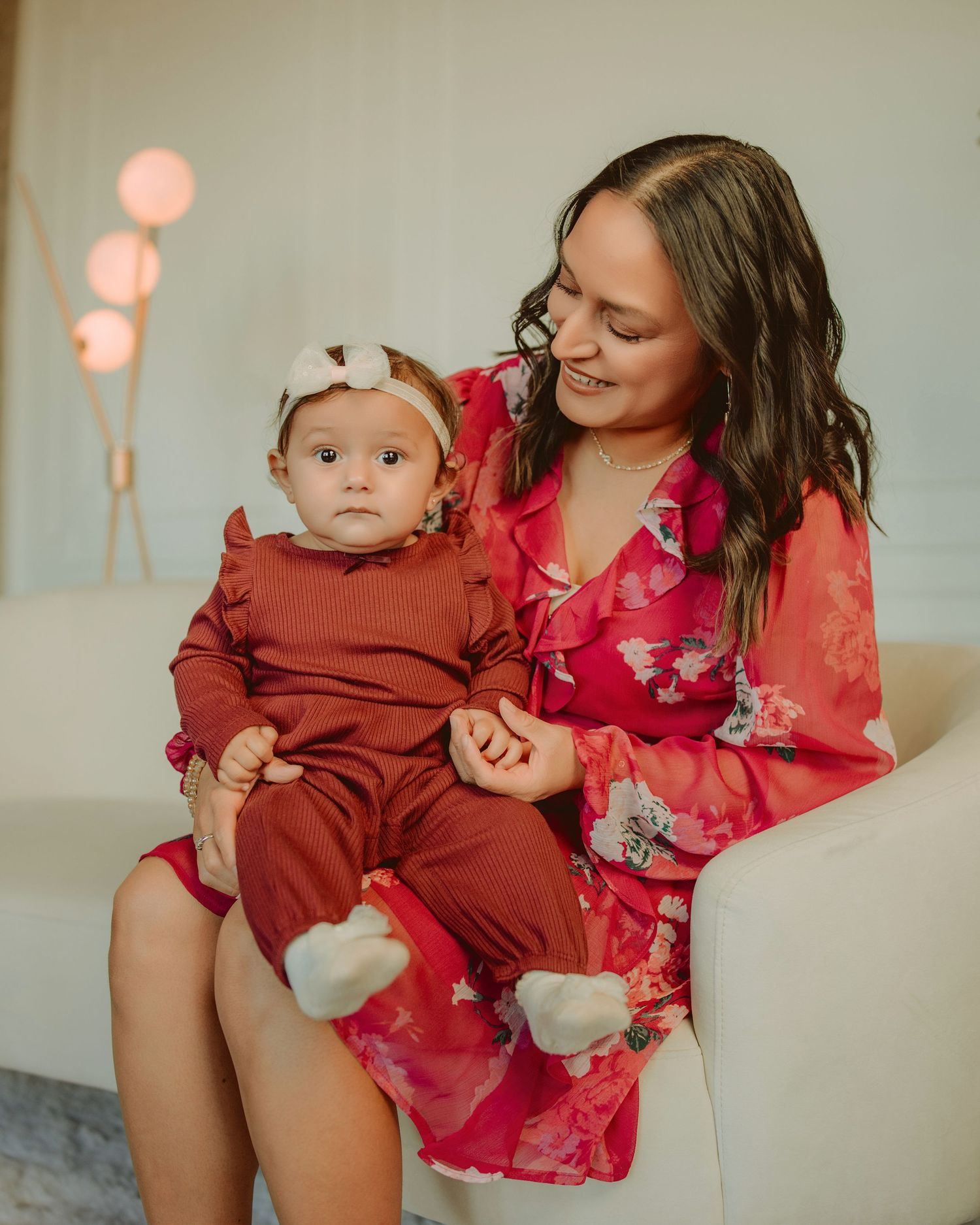 Mother and baby in burgundy clothing pose together on beige background with soft pink lighting.