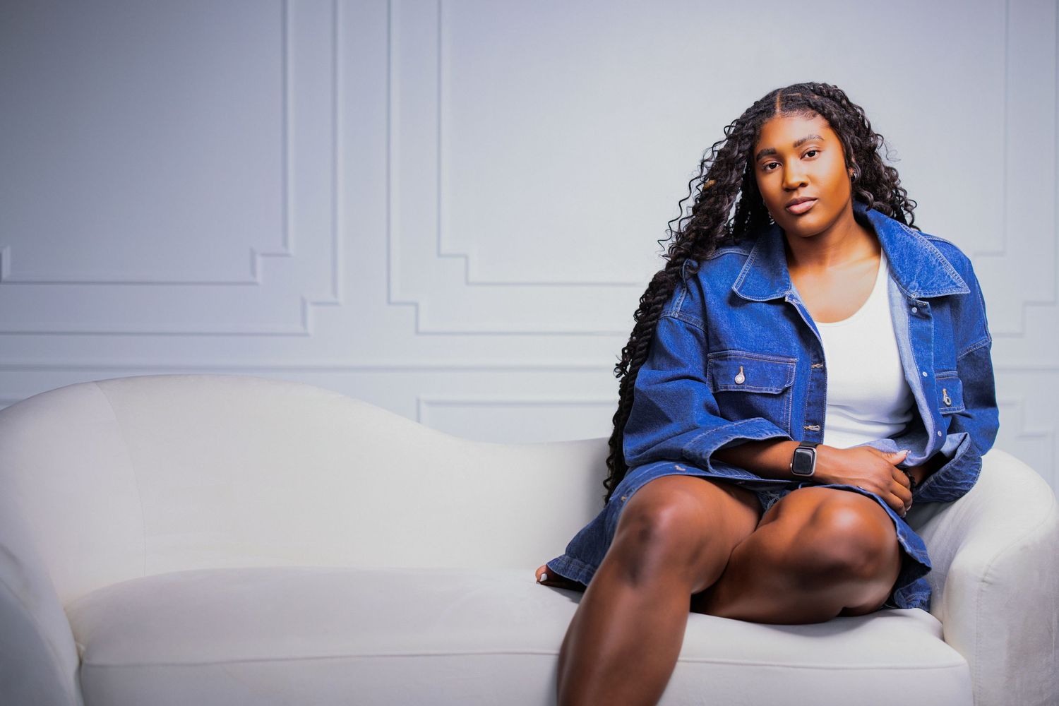 Woman in denim jacket poses casually against white studio wall with architectural details.
