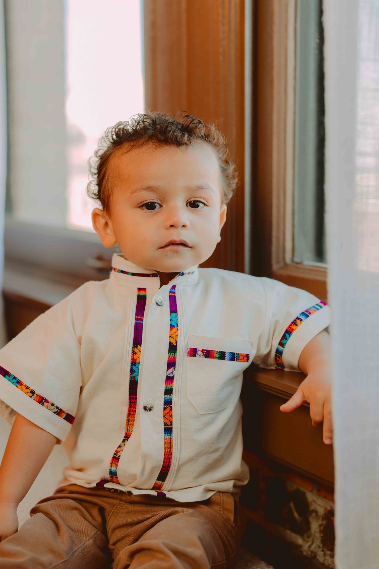 Toddler wearing white outfit with colorful shirt sits near window.