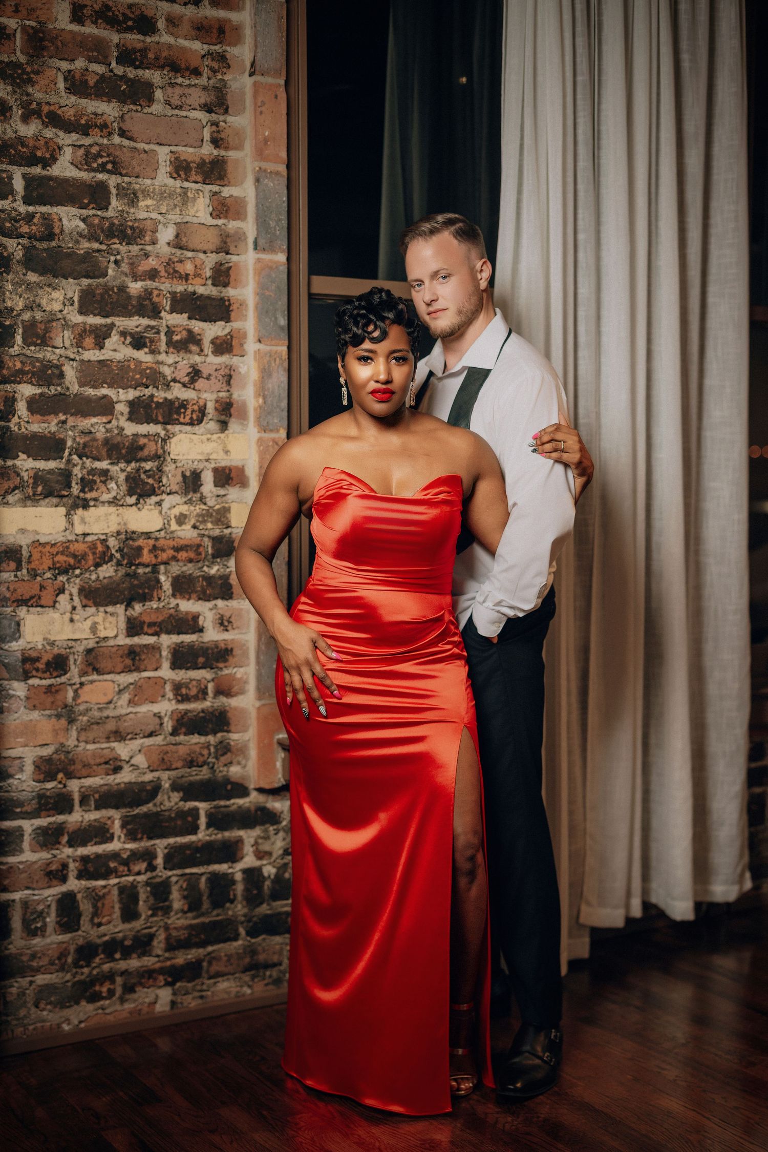 A couple in formal attire poses intimately against a rustic brick wall in a dimly lit room with white curtains.