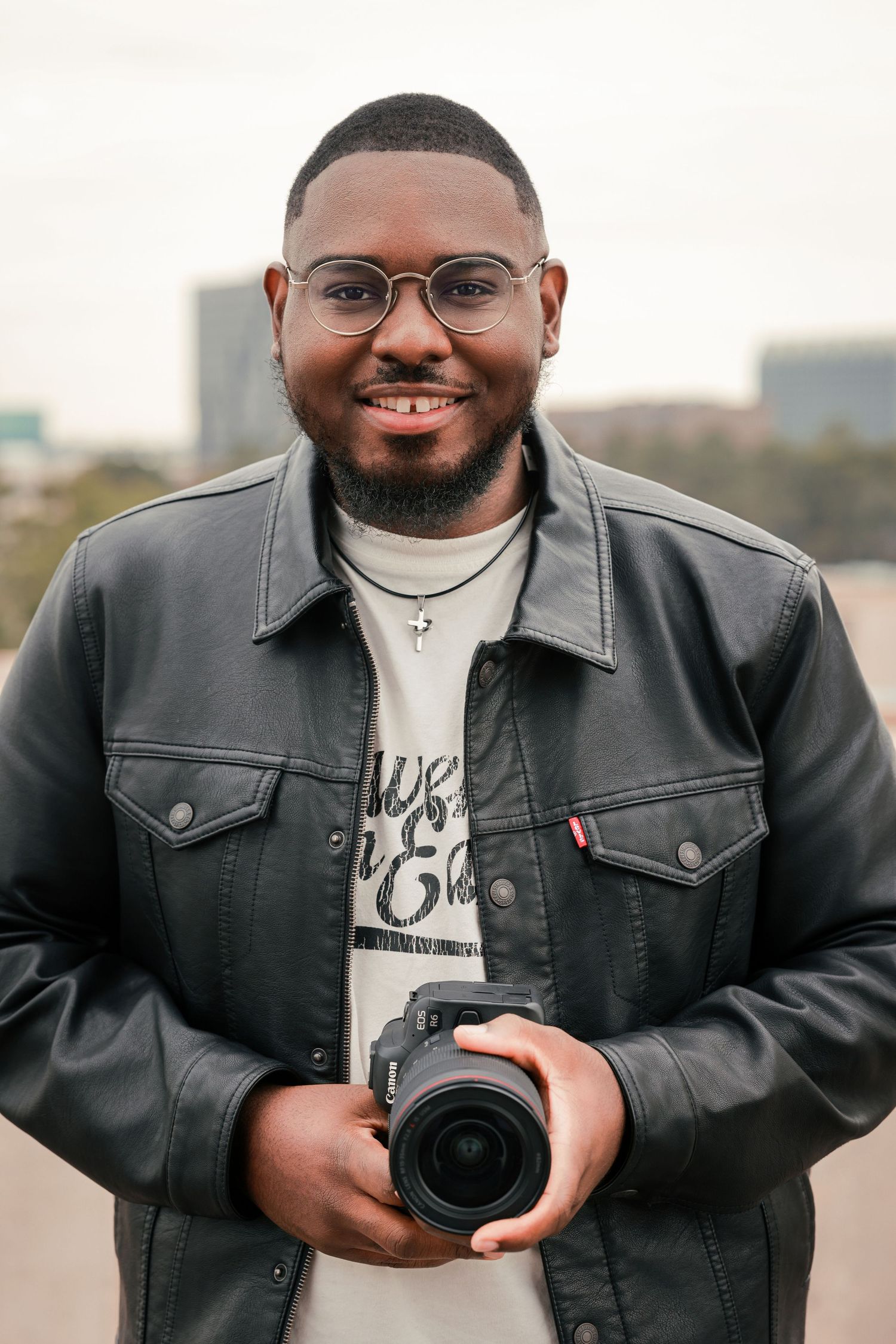 Photographer in black denim jacket holding camera and smiling in outdoor urban setting.