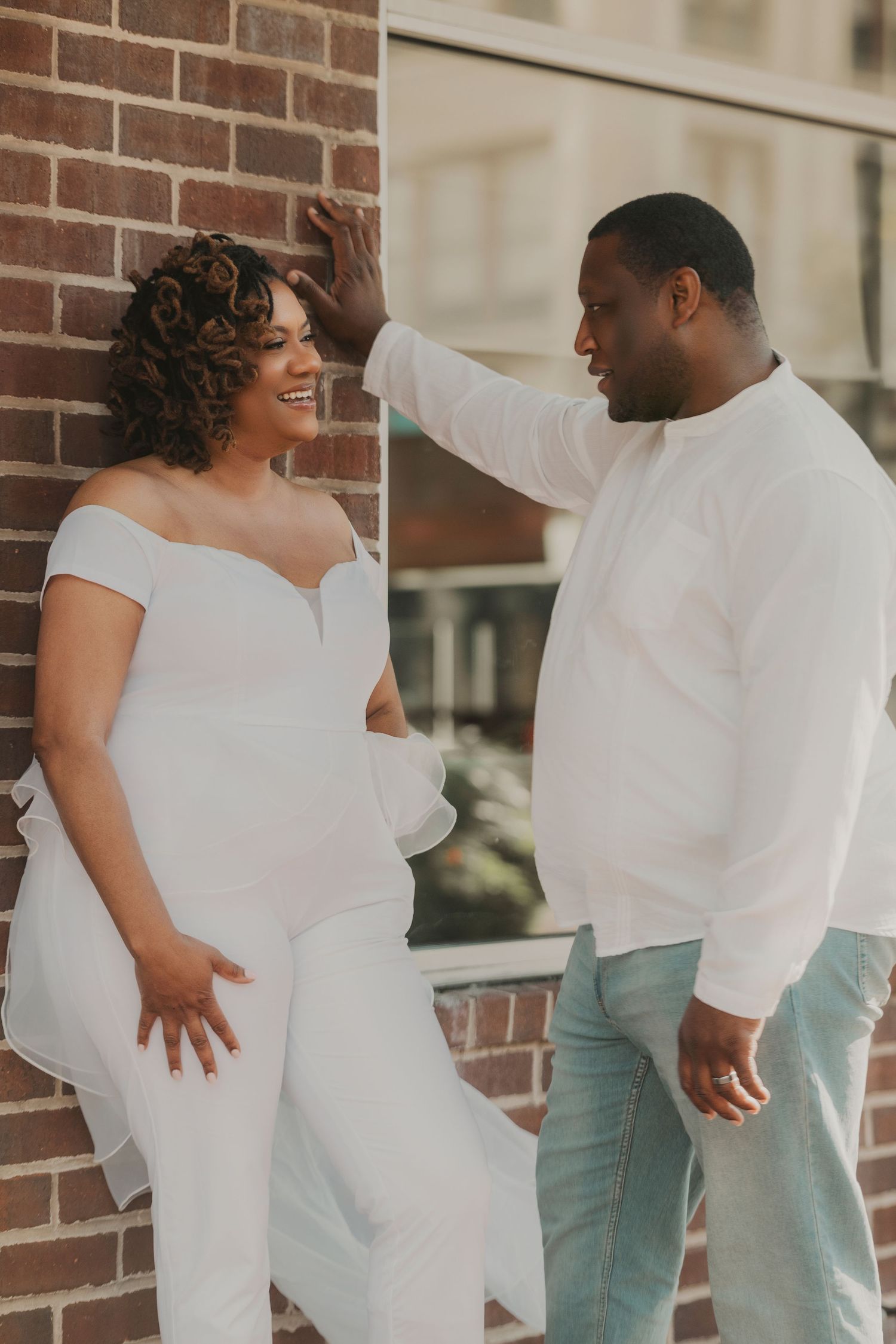 A couple wearing coordinated white outfits poses playfully against a brick wall.