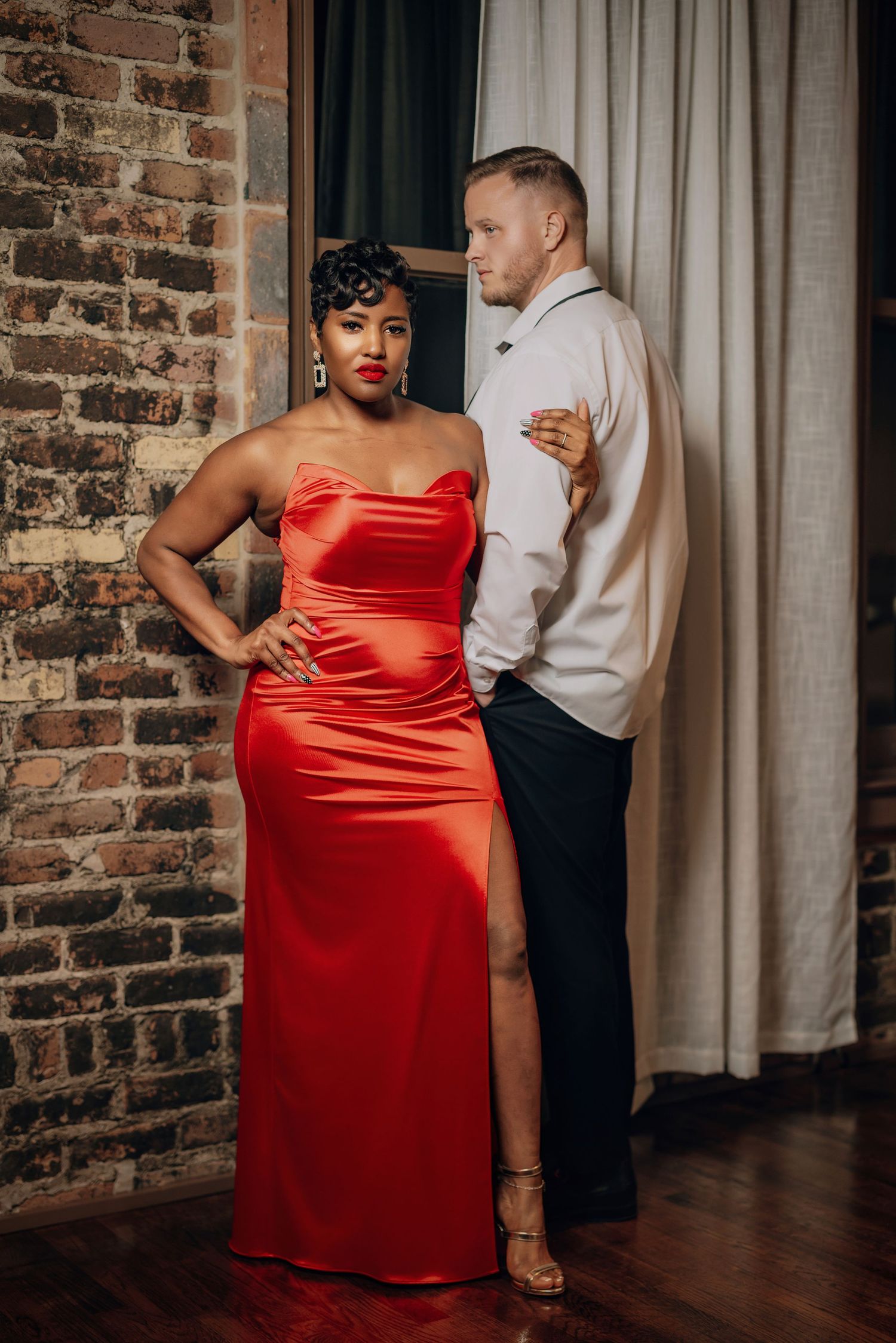 A couple in formal attire dances intimately against a rustic brick wall backdrop, with the woman wearing a red satin dress.