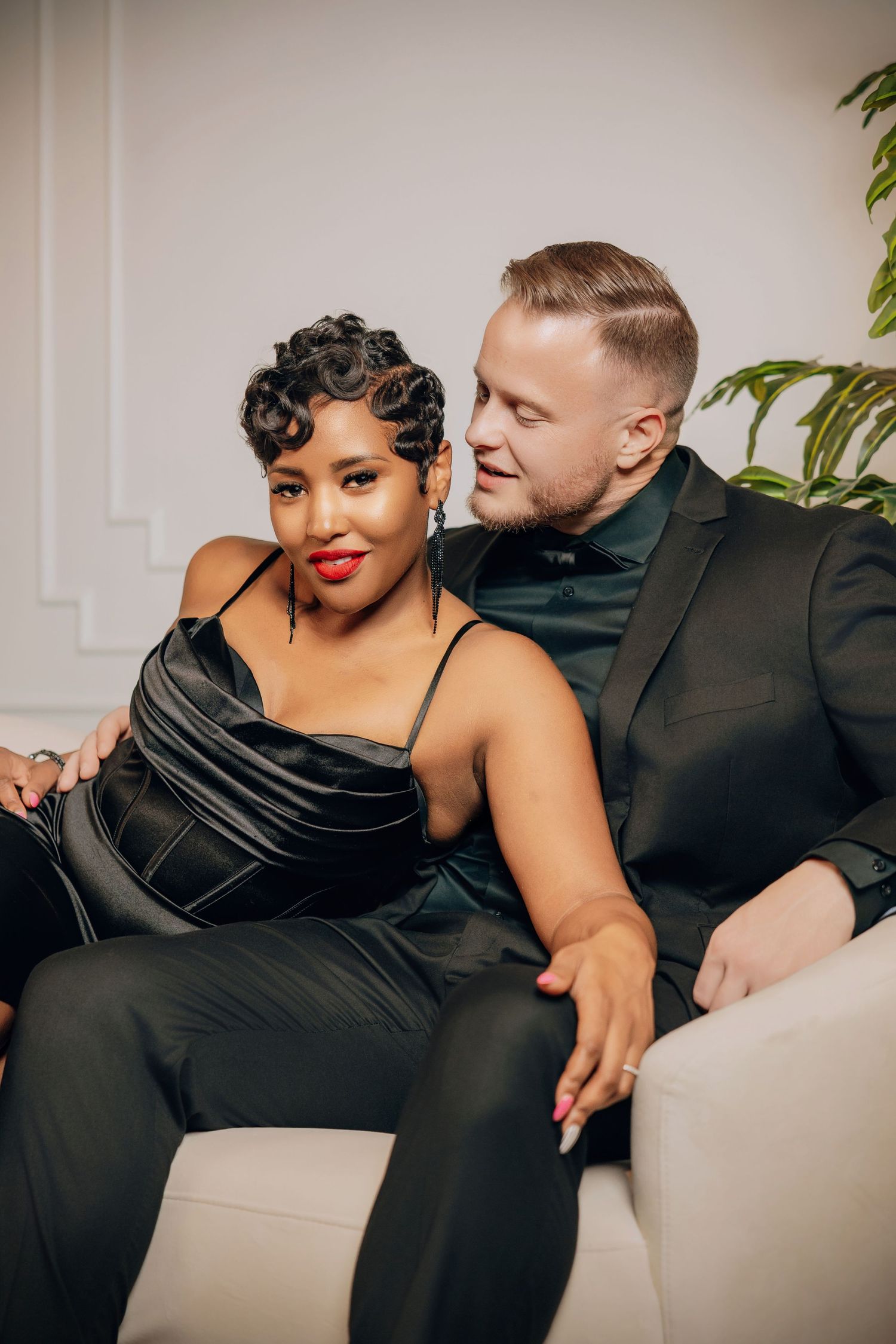 A couple in elegant black attire pose intimately on a white couch in a modern interior setting.