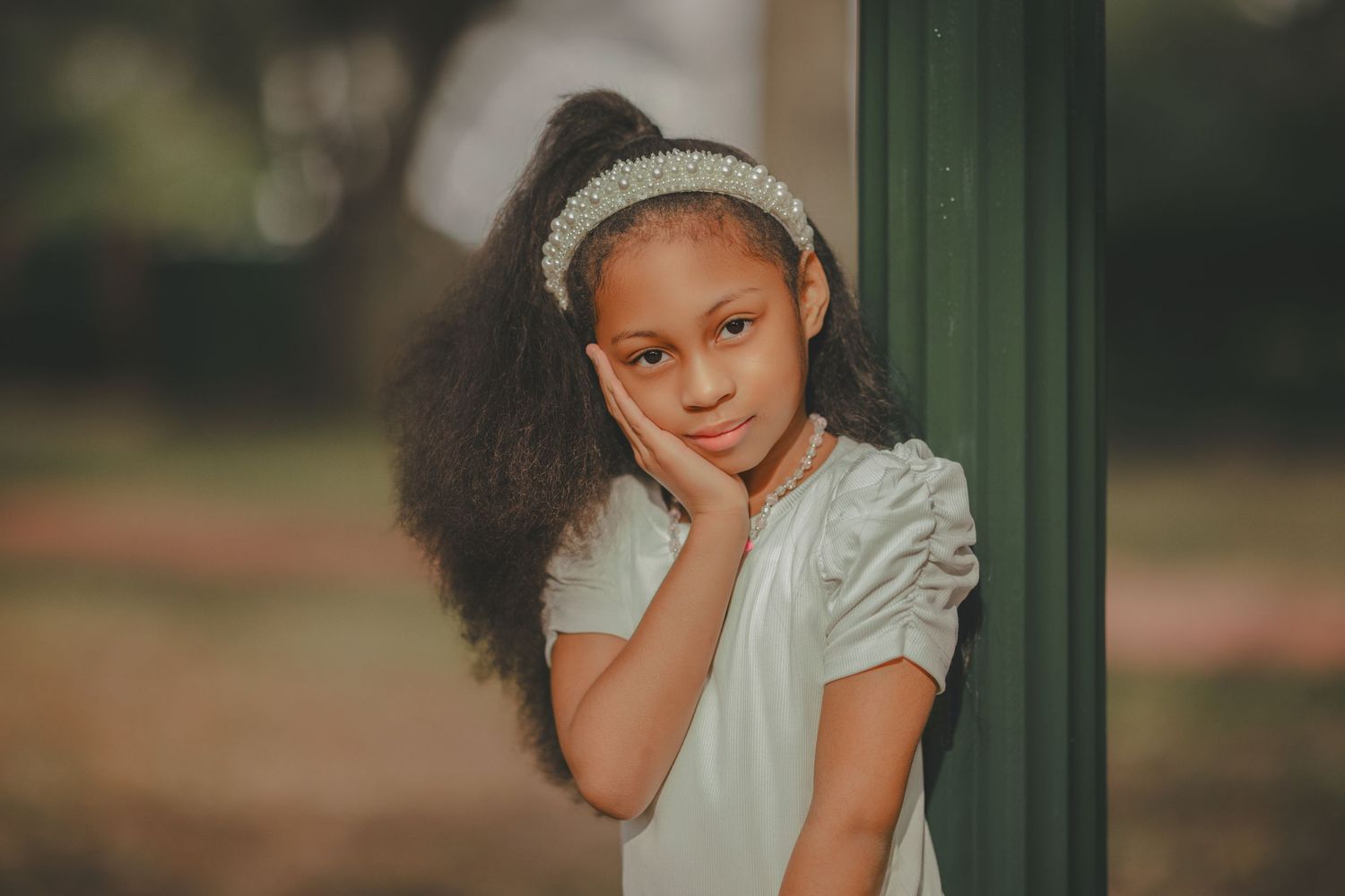 A child in a white dress and beanie poses outdoors against a blurred green background.