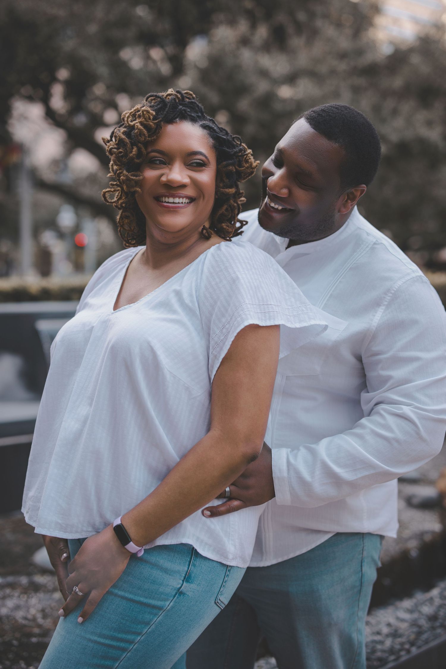 A couple wearing matching white tops and blue jeans poses playfully outdoors.