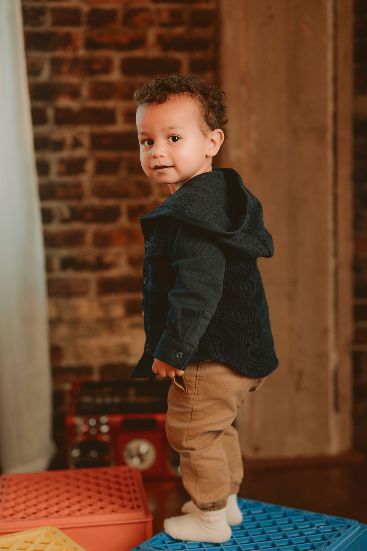 A toddler in a black hoodie and khaki pants stands on colorful play tiles against a brick wall background.