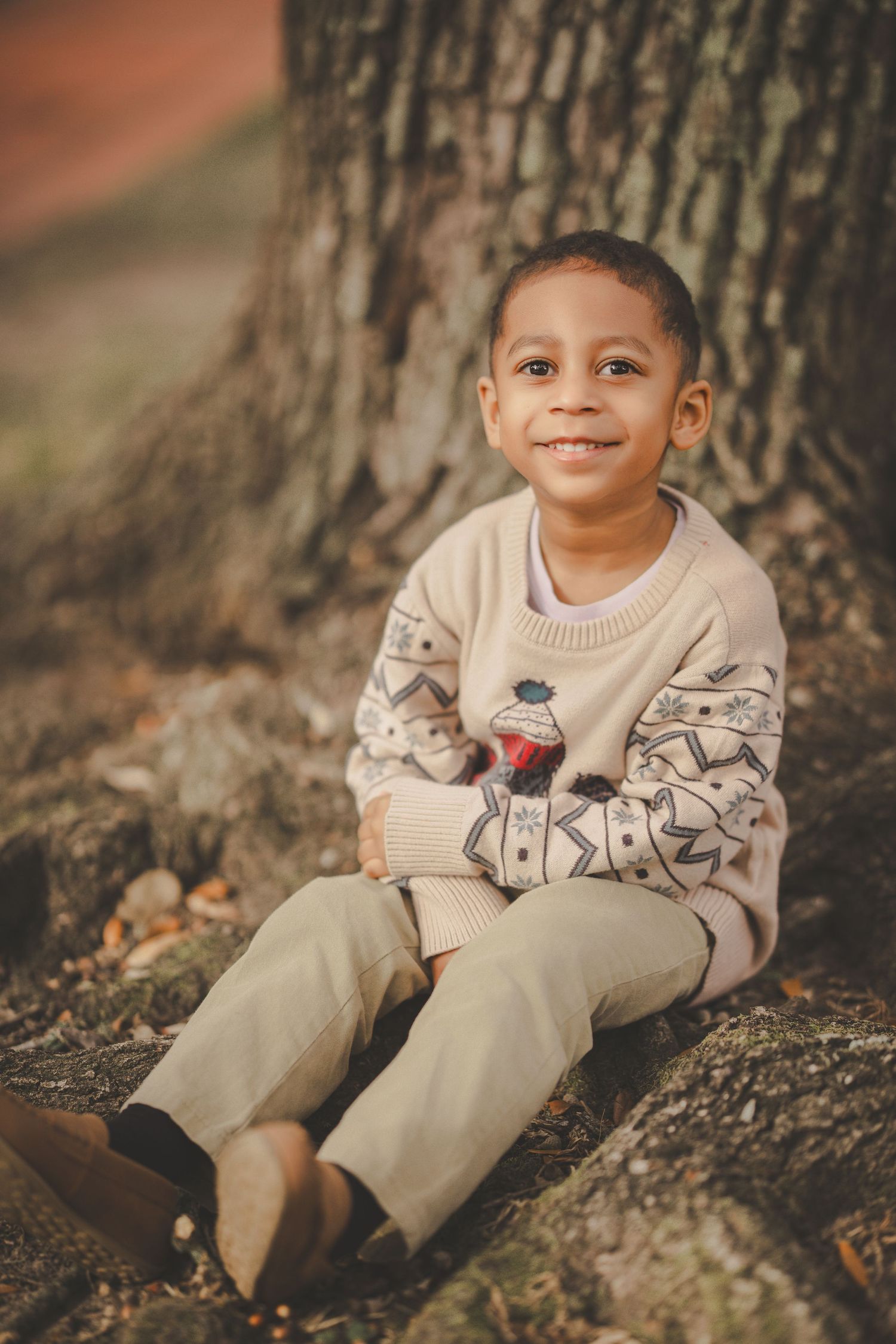 A young child sits comfortably against a tree trunk wearing a cozy patterned sweater.