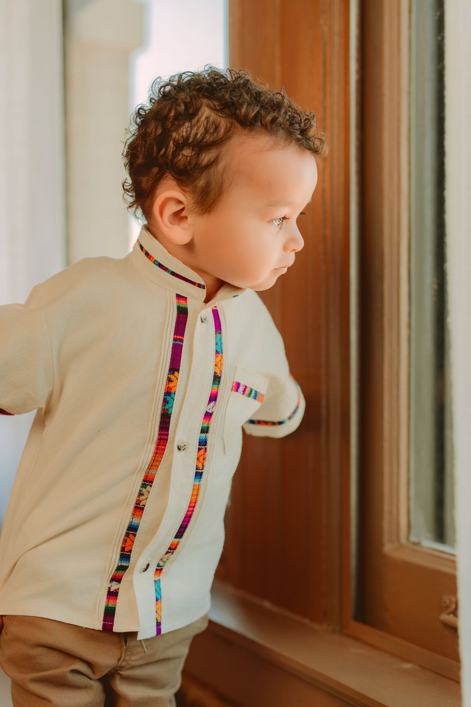 A small child in a beige outfit with colorful striped accents stands near a window in a series of connected photos.
