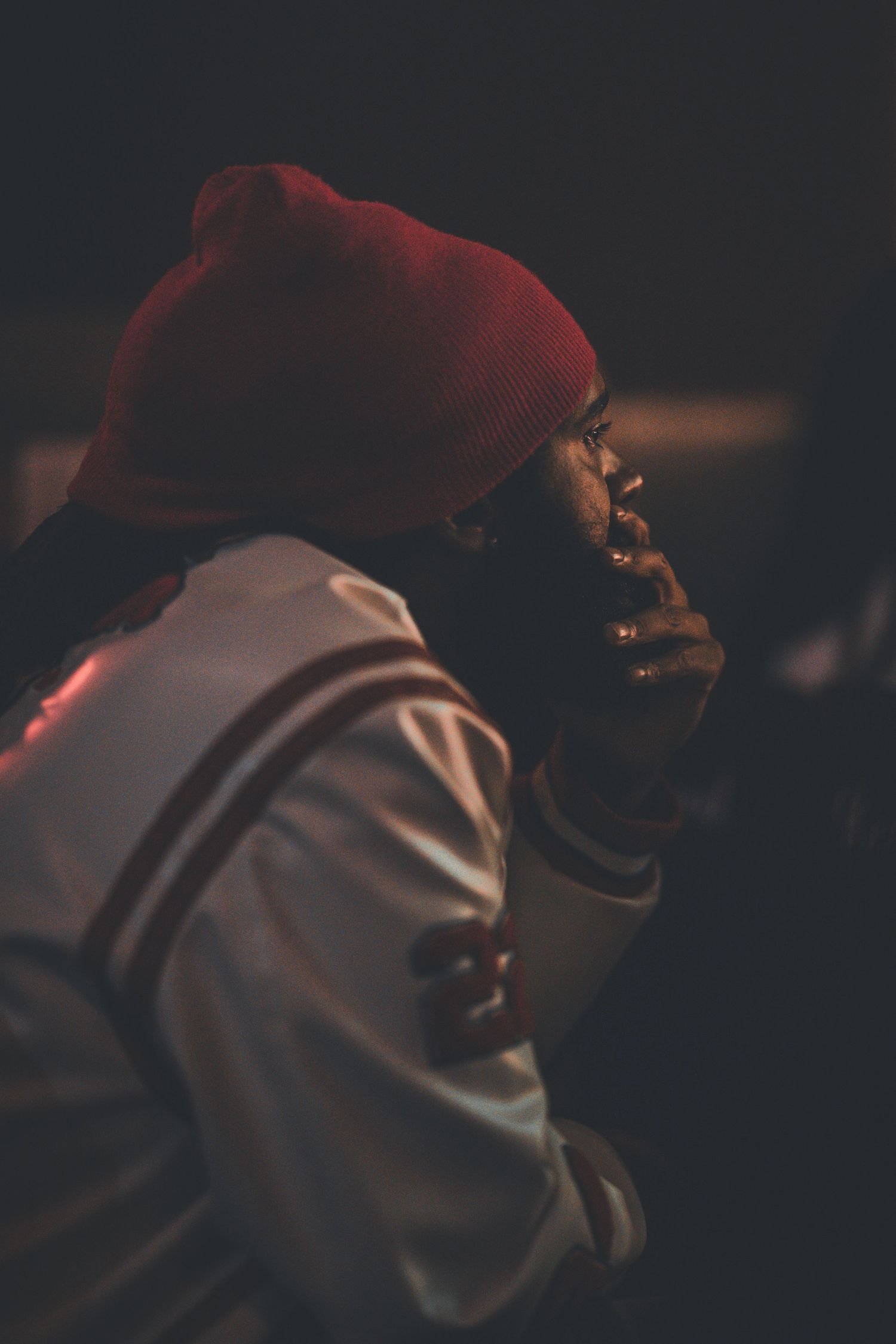A contemplative figure in a red beanie and light jacket sits in dim lighting.