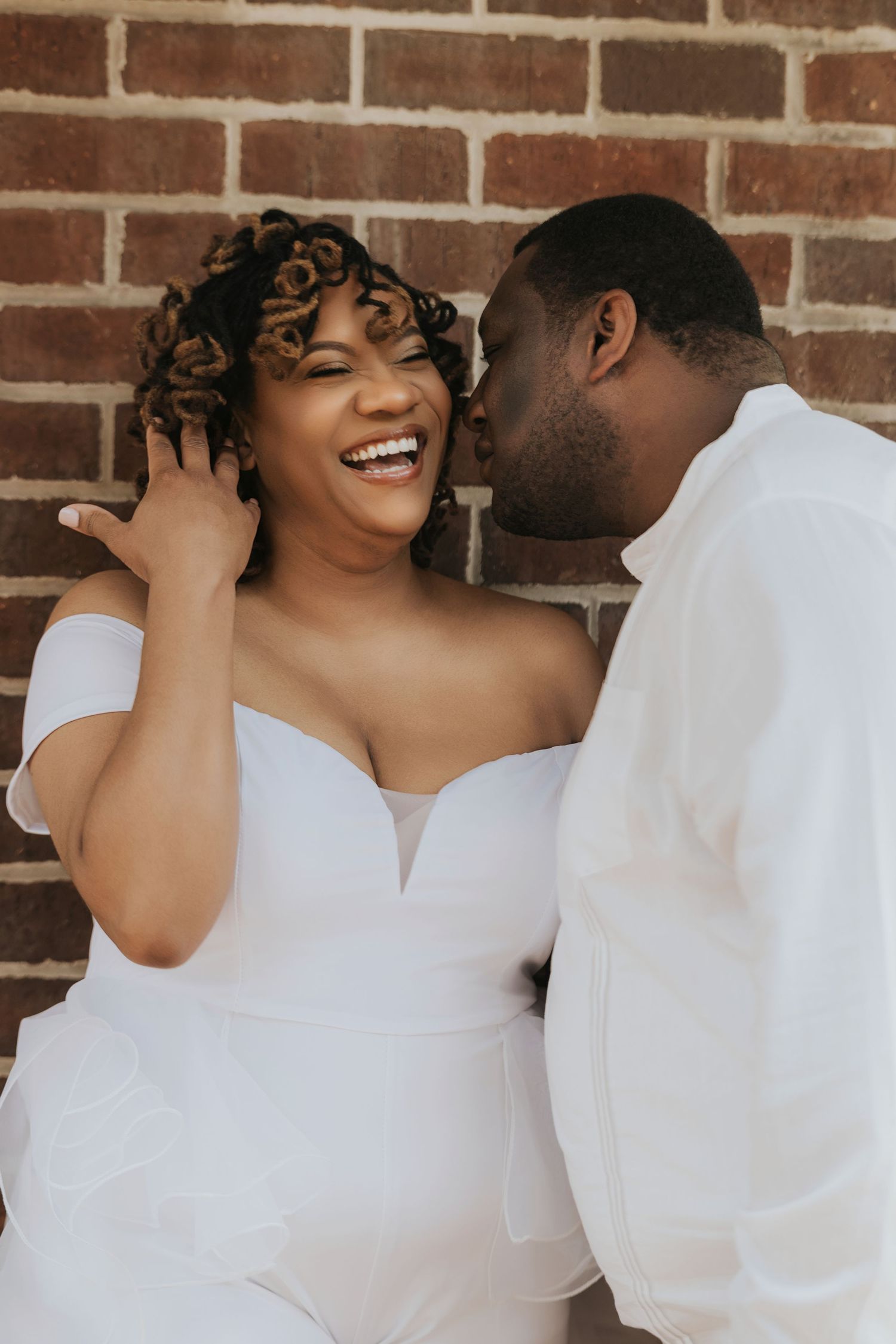 A couple in white outfits shares loving glances against a brick wall background.