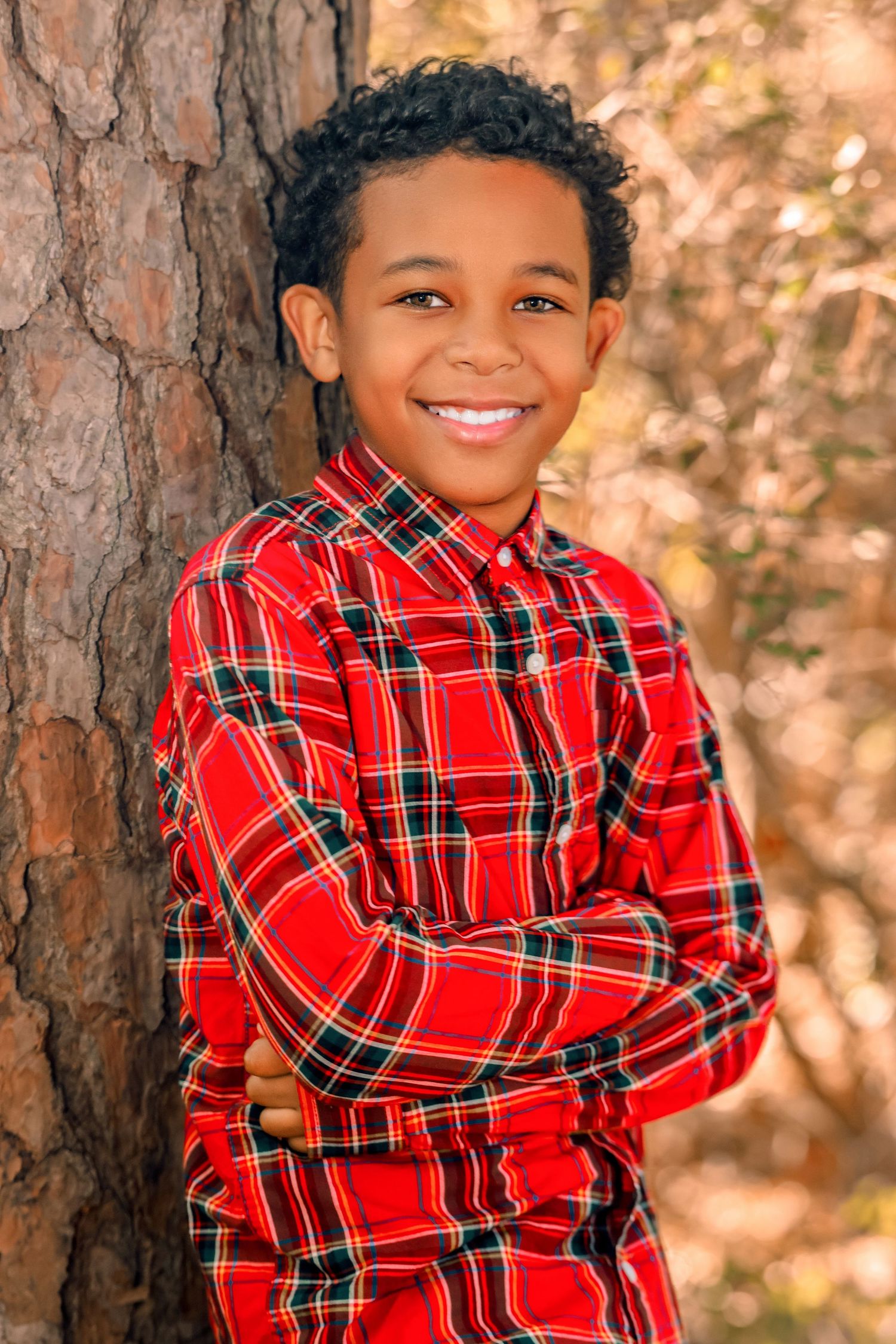 Child wearing red plaid shirt poses playfully against autumn trees and warm lighting.