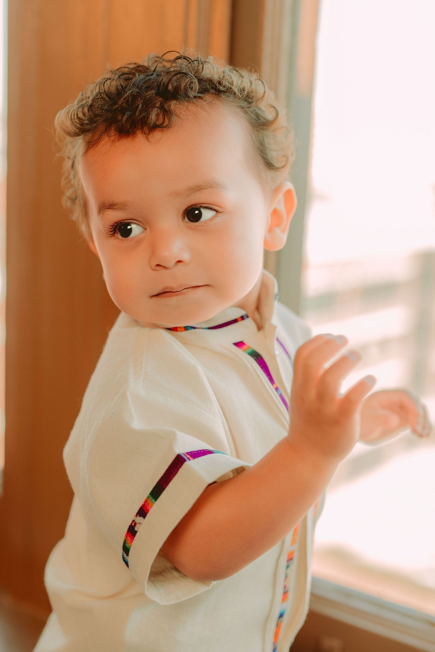 Young child in white polo shirt with colorful stripes stands by window in warm lighting.