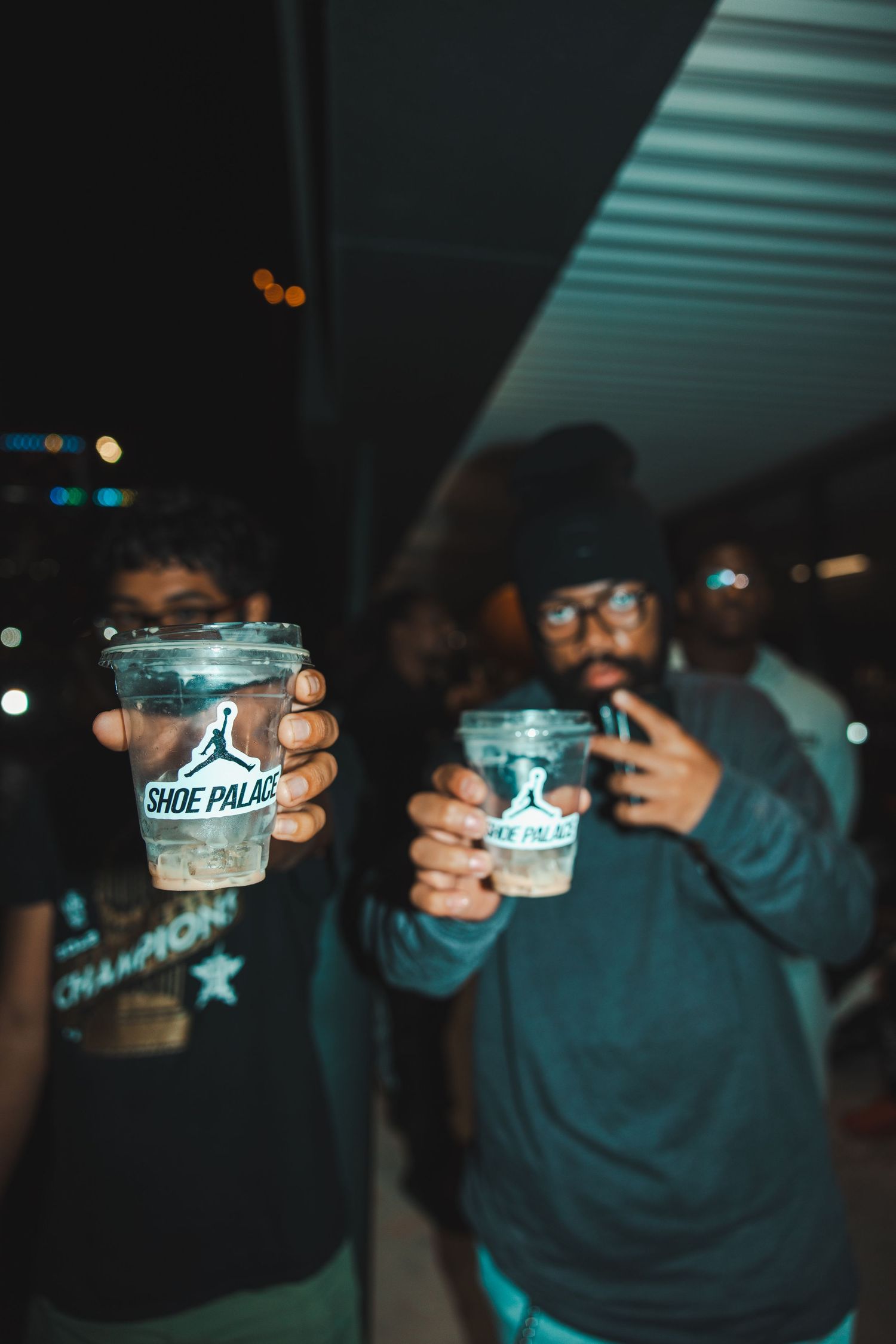 Several hands hold up clear plastic cups in a toast under dark lighting with bokeh effects.