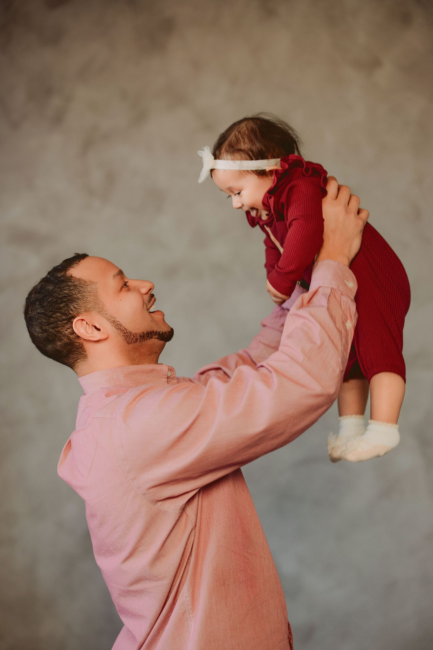 A parent in a pink sweater lifts a small child wearing red, sharing a joyful moment against a neutral background.