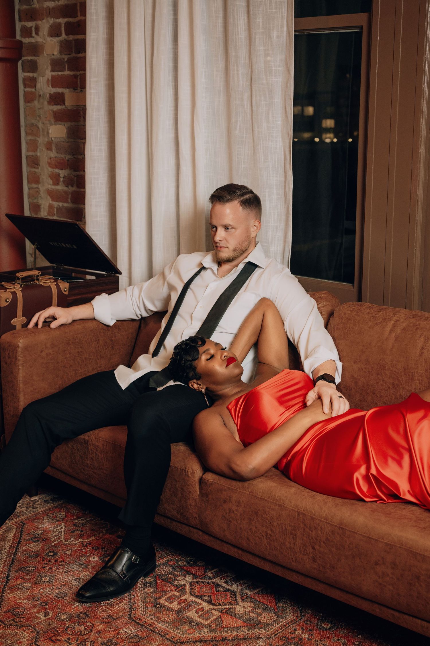 Romantic couple lounges on brown leather couch, with one in formal attire and the other in red satin dress.