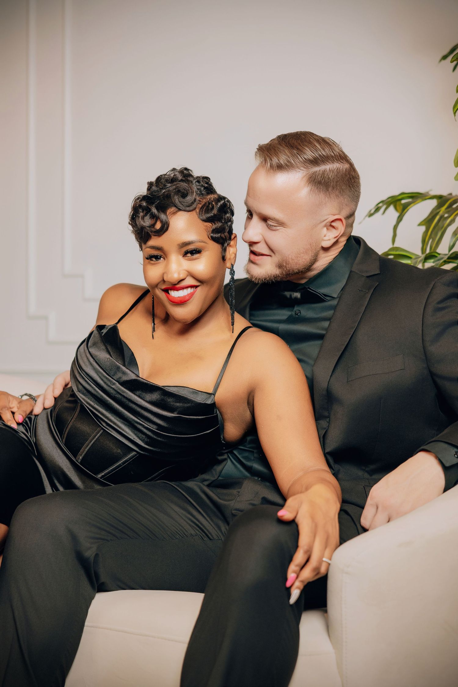 A couple in black attire shares an intimate moment while sitting together on a light colored couch.