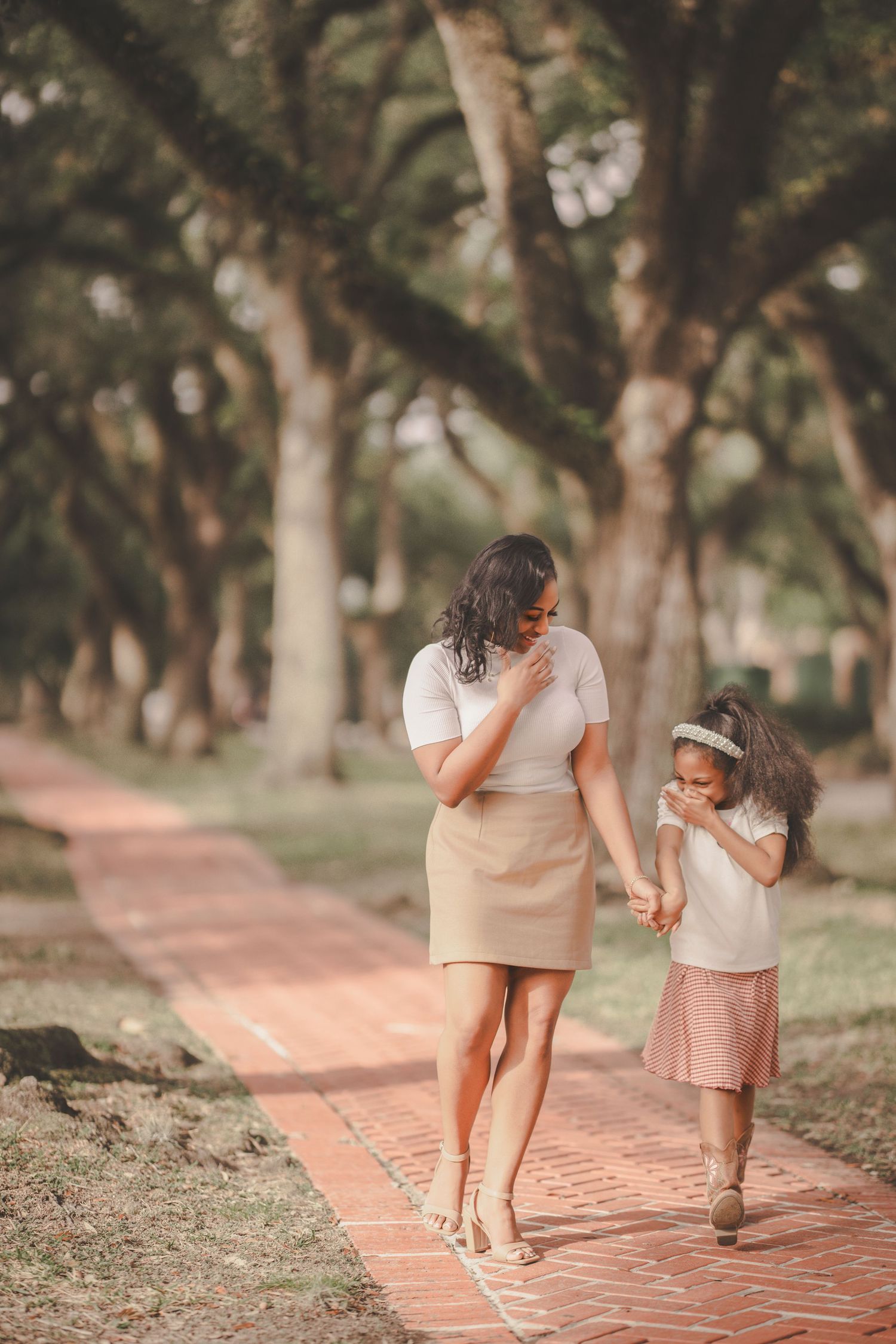 Mother and child walking hand in hand along a brick pathway surrounded by trees in a park setting.
