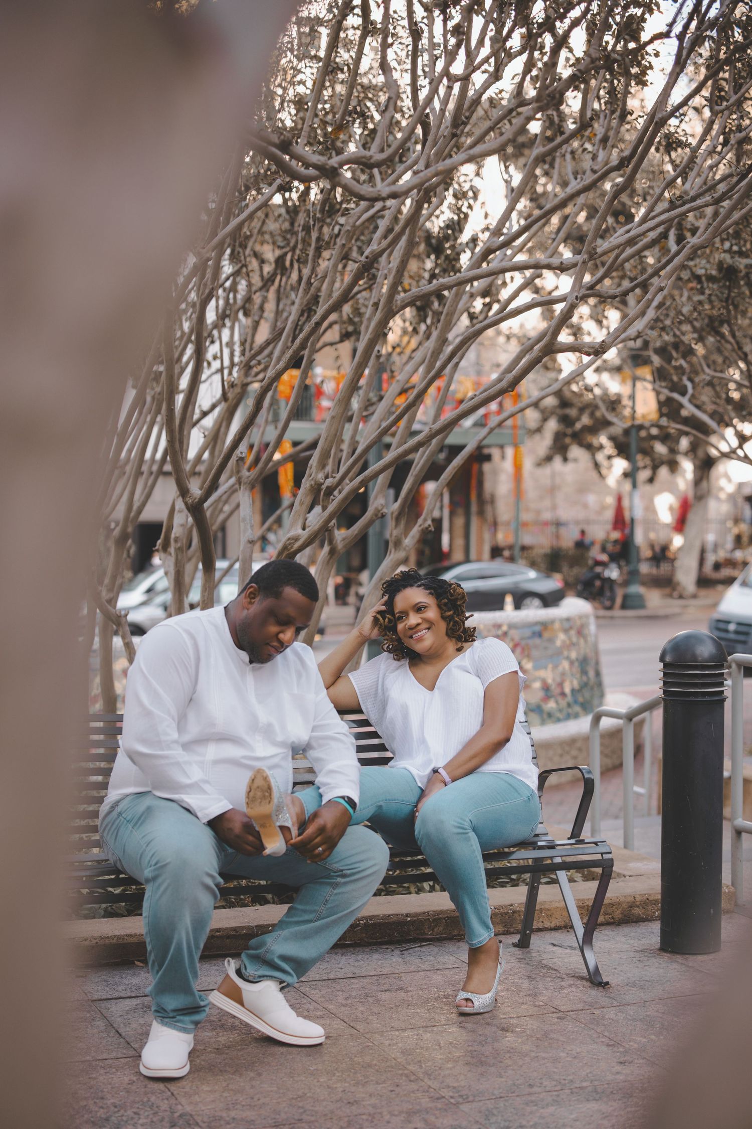 A couple in white tops and blue jeans sits together on a bench in an outdoor setting.