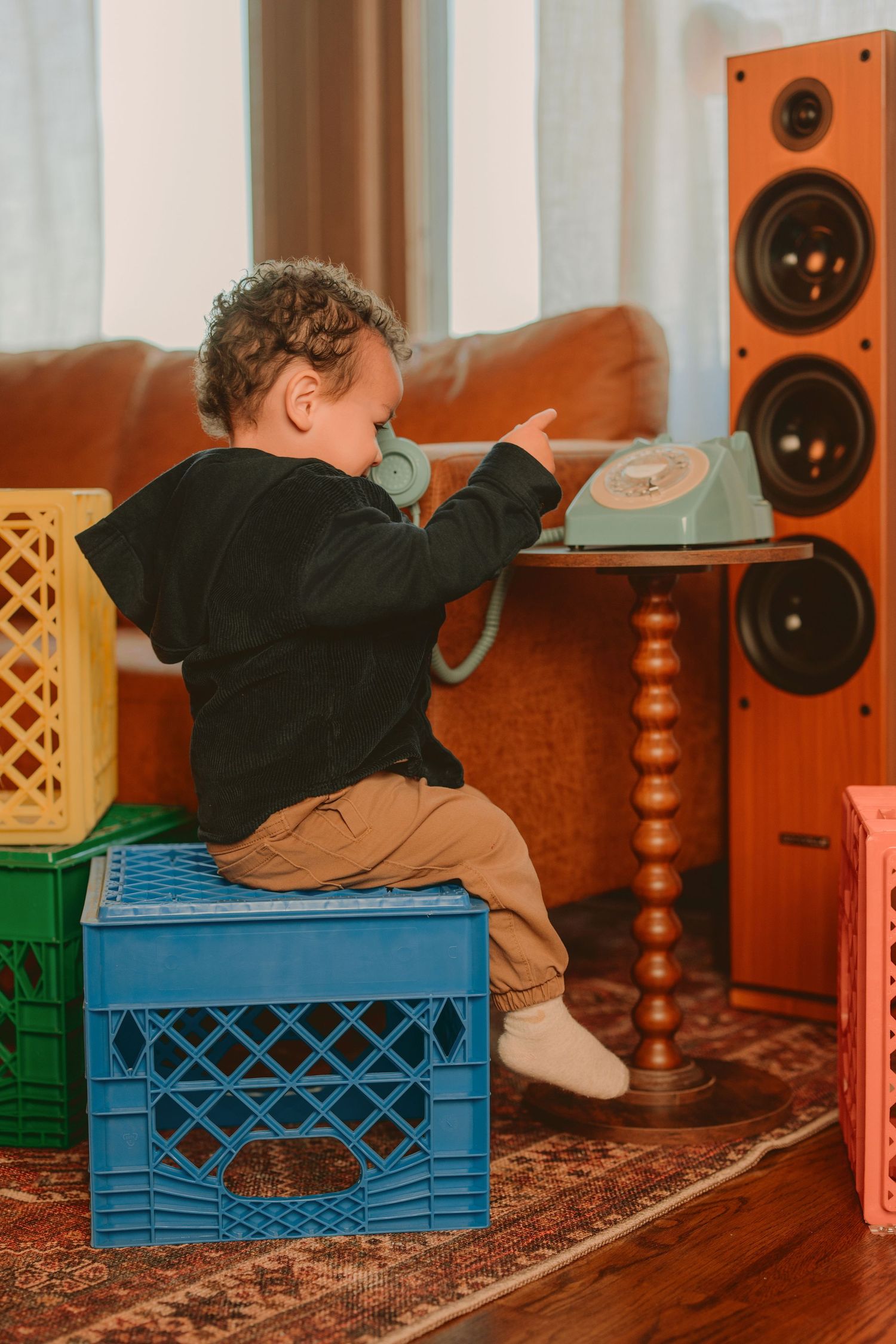 A child lies on a blue milk crate while listening to music through headphones on a patterned rug.