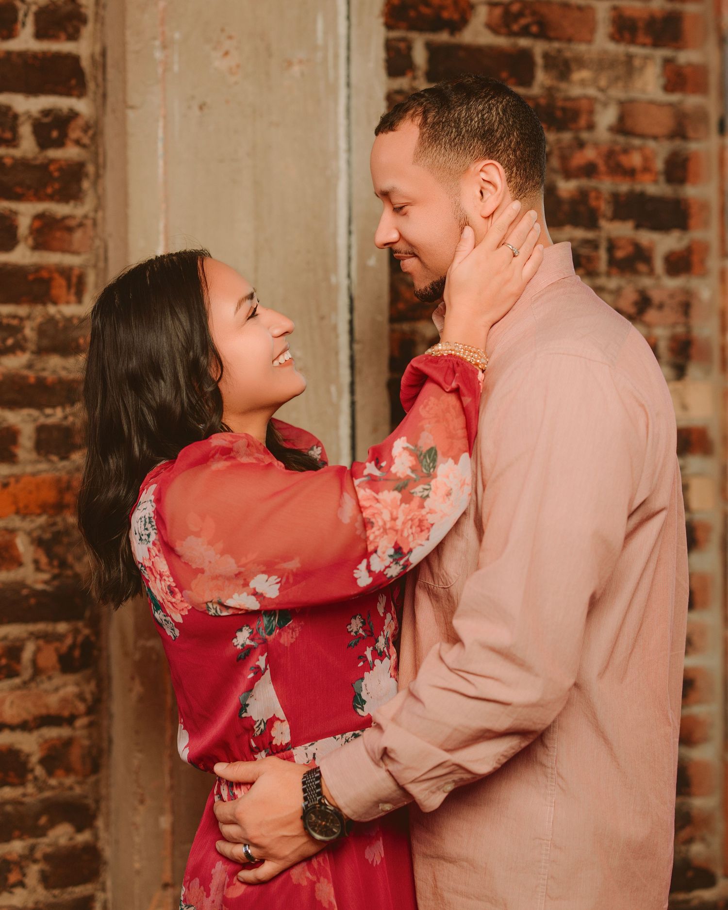 A couple embraces and laughs together against a rustic brick wall, with one person wearing a vibrant red floral dress.